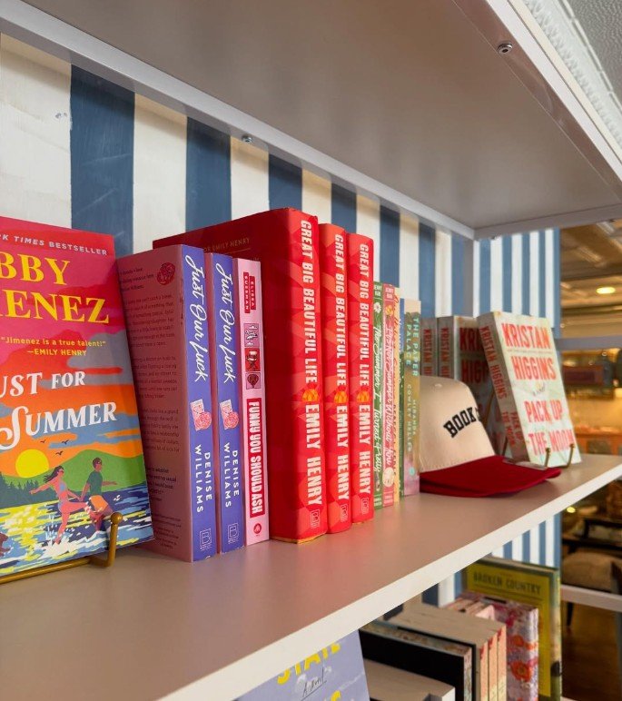 Books on a white and blue striped bookshelf, including 'Baby Sitter' by Emily Henry, 'Just for Summer' by Jenny Han, and others, along with a white cap with the word 'BOOK' and a red brim.