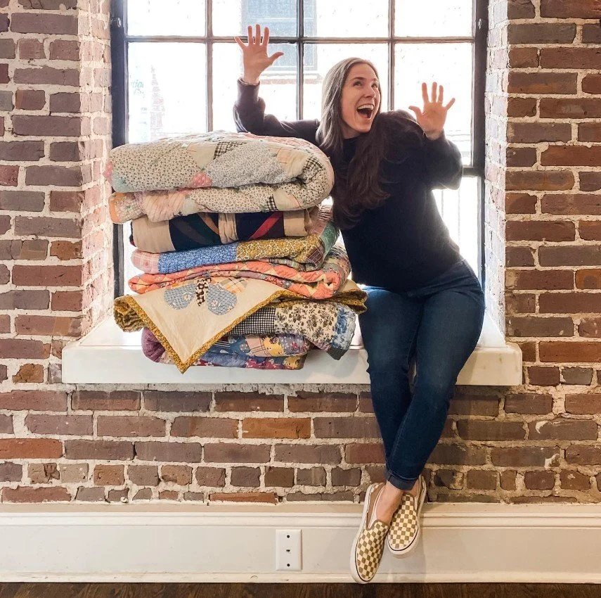 A woman sitting on a windowsill with a stack of colorful quilts, smiling and raising her hands, wearing checkered slip-on shoes, in front of a brick wall.