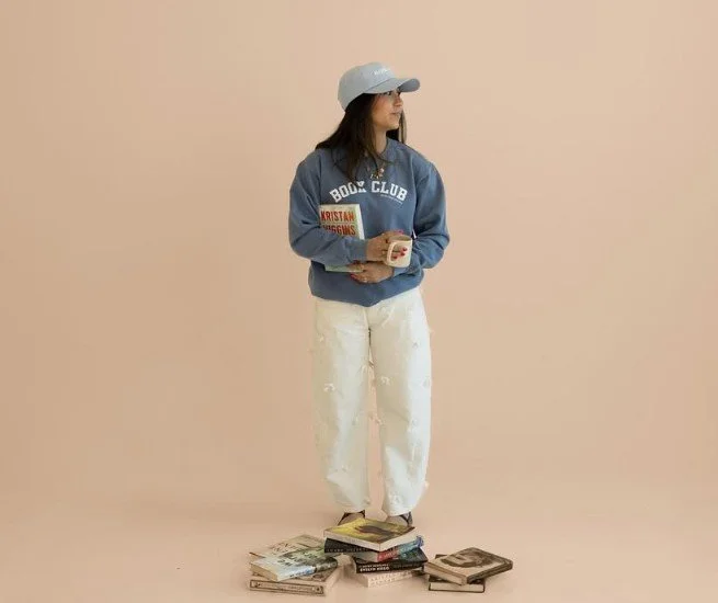 A woman standing on a stack of books reading a book, wearing a blue hoodie, white pants, and a gray cap, against a plain beige background.