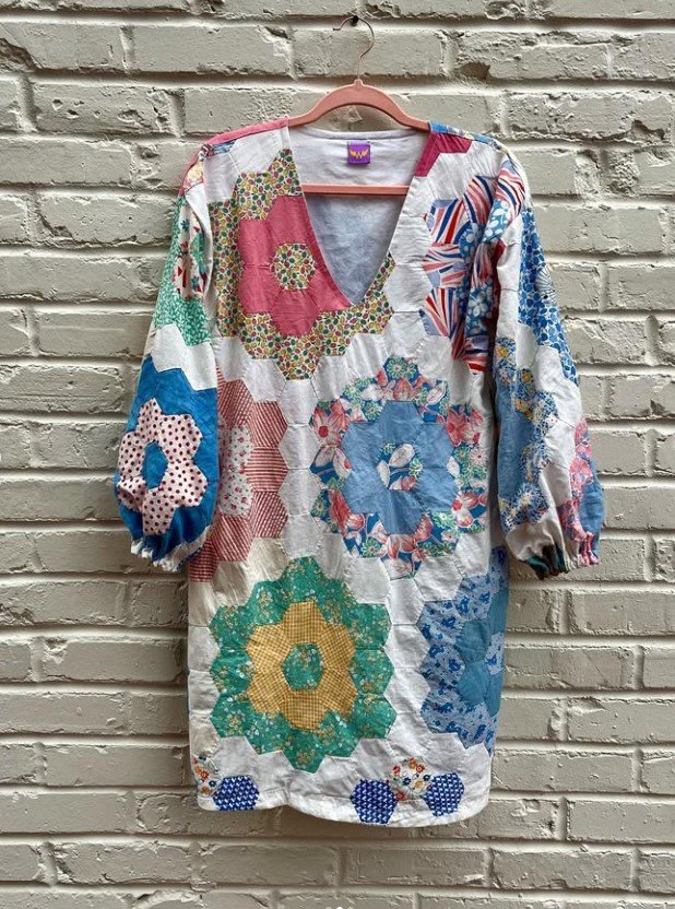 A colorful patchwork dress with a V neckline and three-quarter length, puffed sleeves hanging on a pink hanger against a white brick wall.