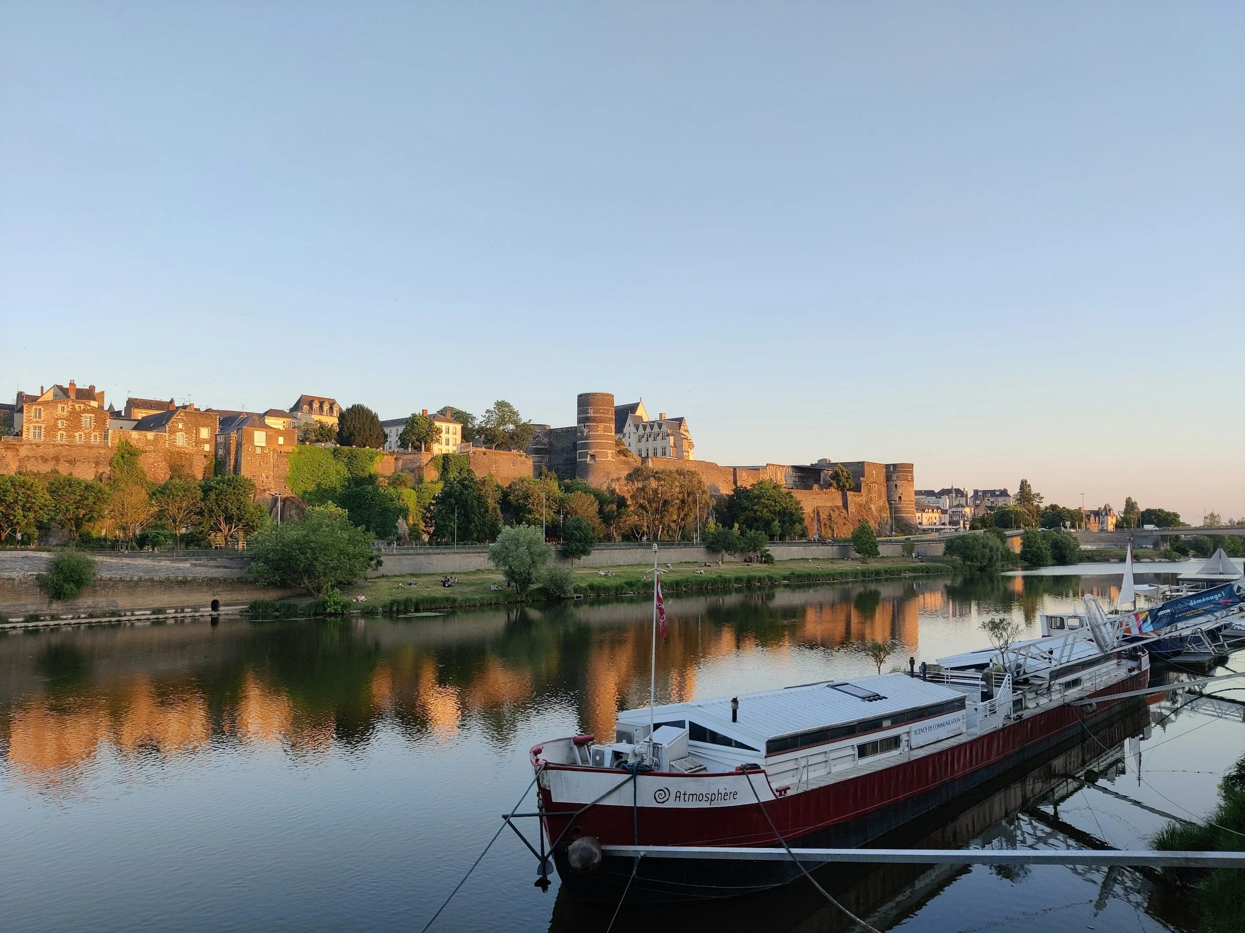 Vue d'une rivière avec un bateau à quai, un château et des maisons anciennes en arrière-plan, sous un ciel clair.