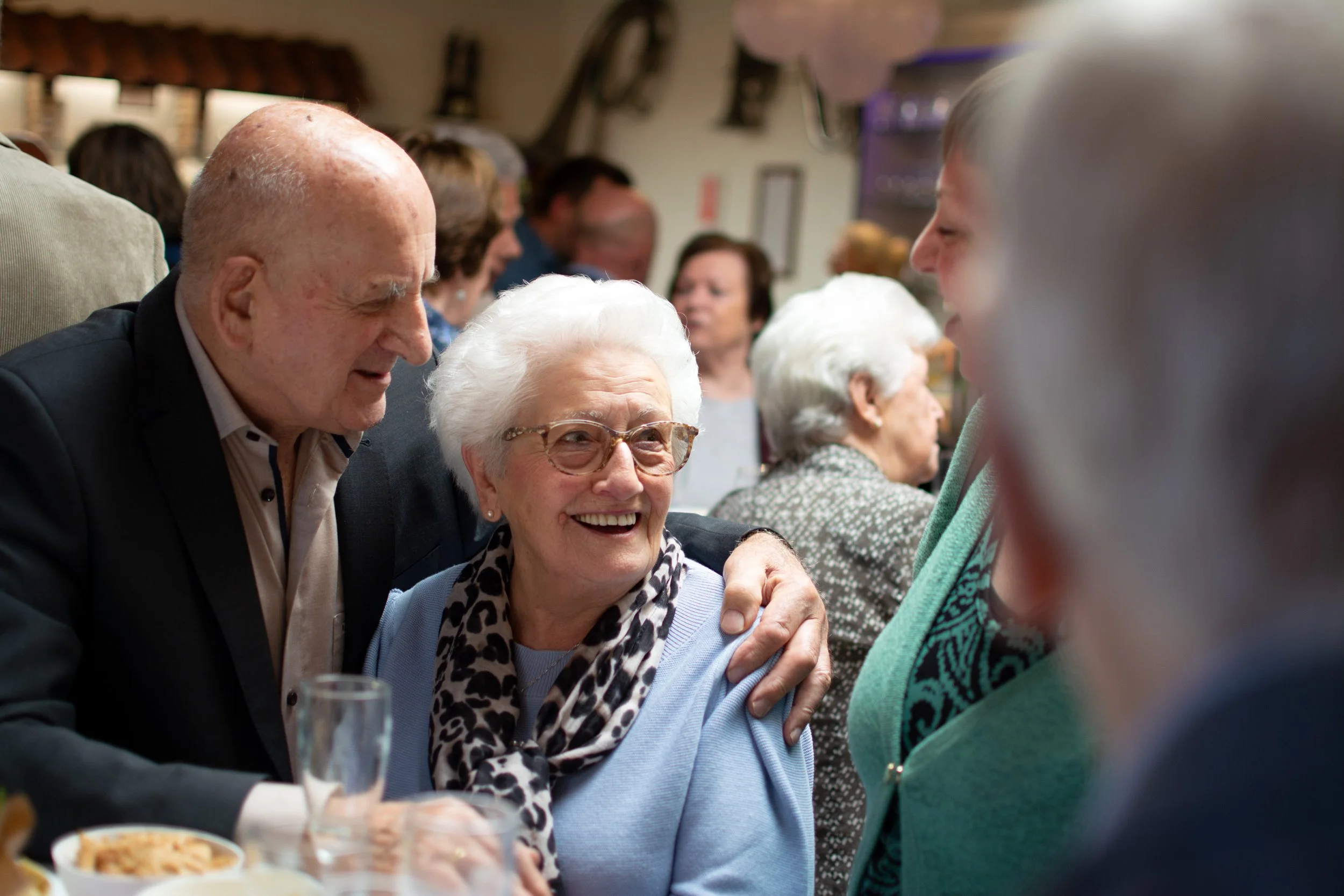 An elderly woman with white hair, glasses, and a leopard print scarf is smiling and being greeted by an elderly man at a social gathering, surrounded by other people.