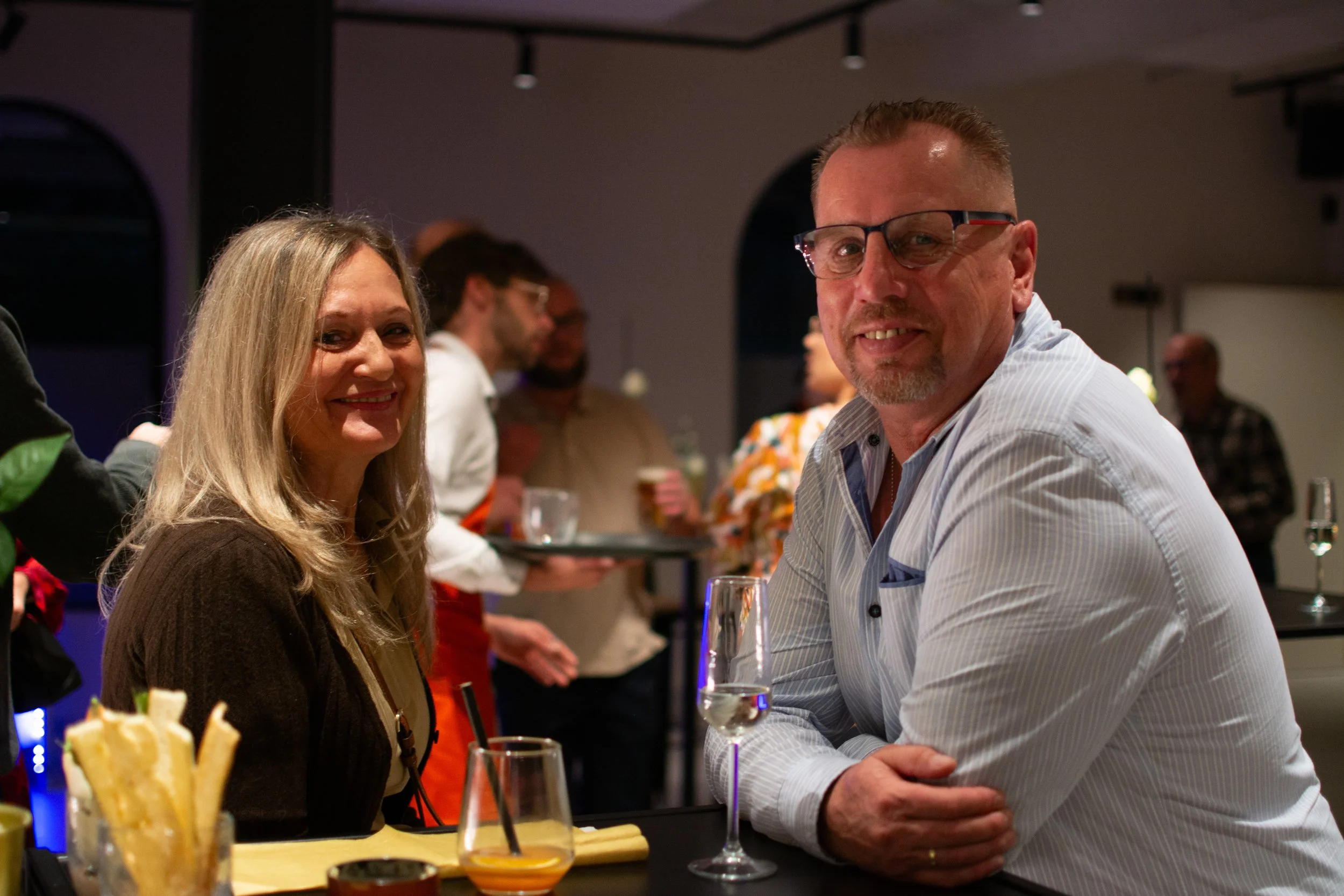 Two people, a woman with blonde hair and a man with glasses, smiling at a social gathering or party. They are seated at a table with drinks and snacks, with other guests in the background.