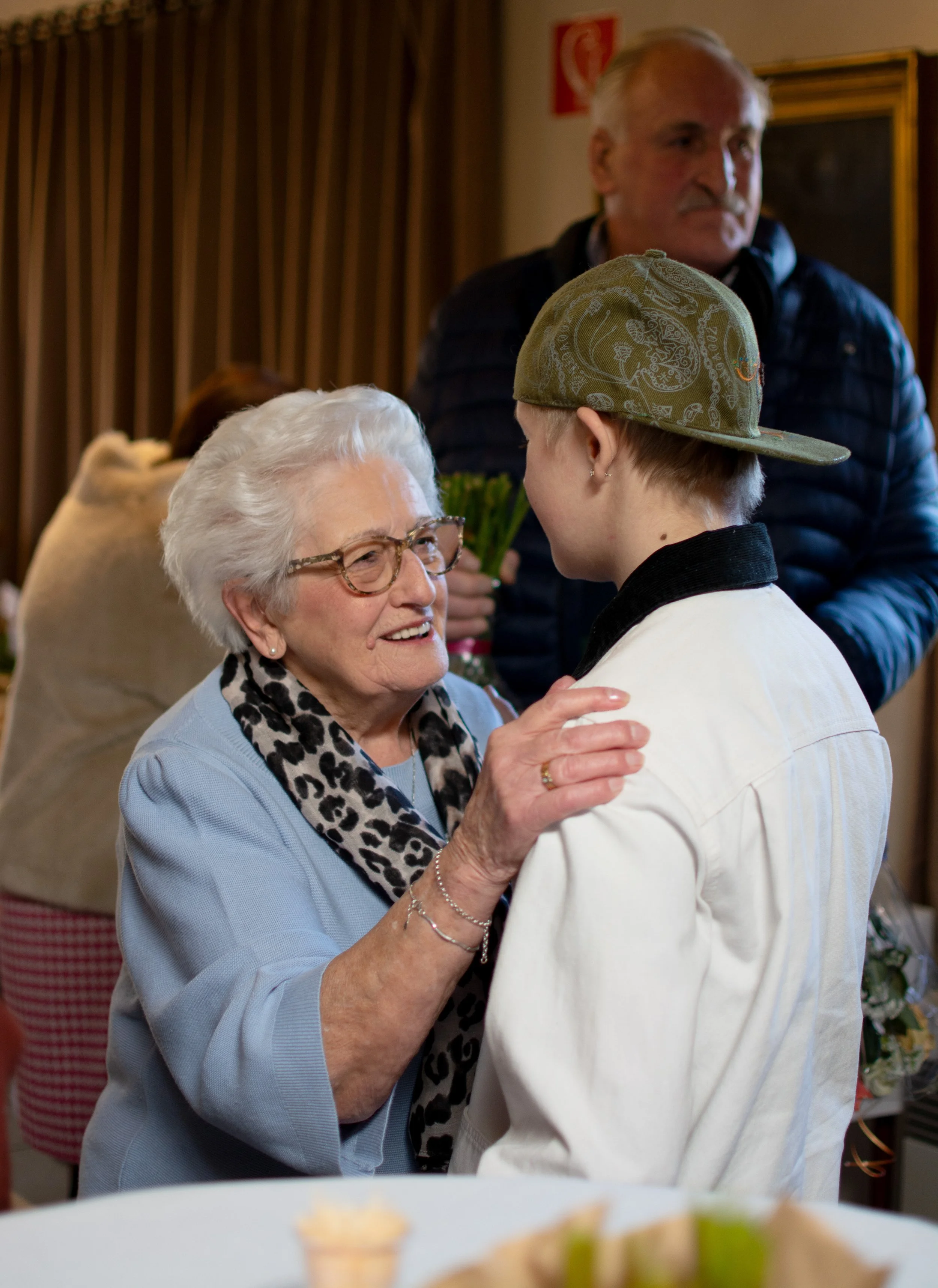 An elderly woman with glasses and a leopard print scarf smiling at a young man wearing a whites blazer and a green cap.