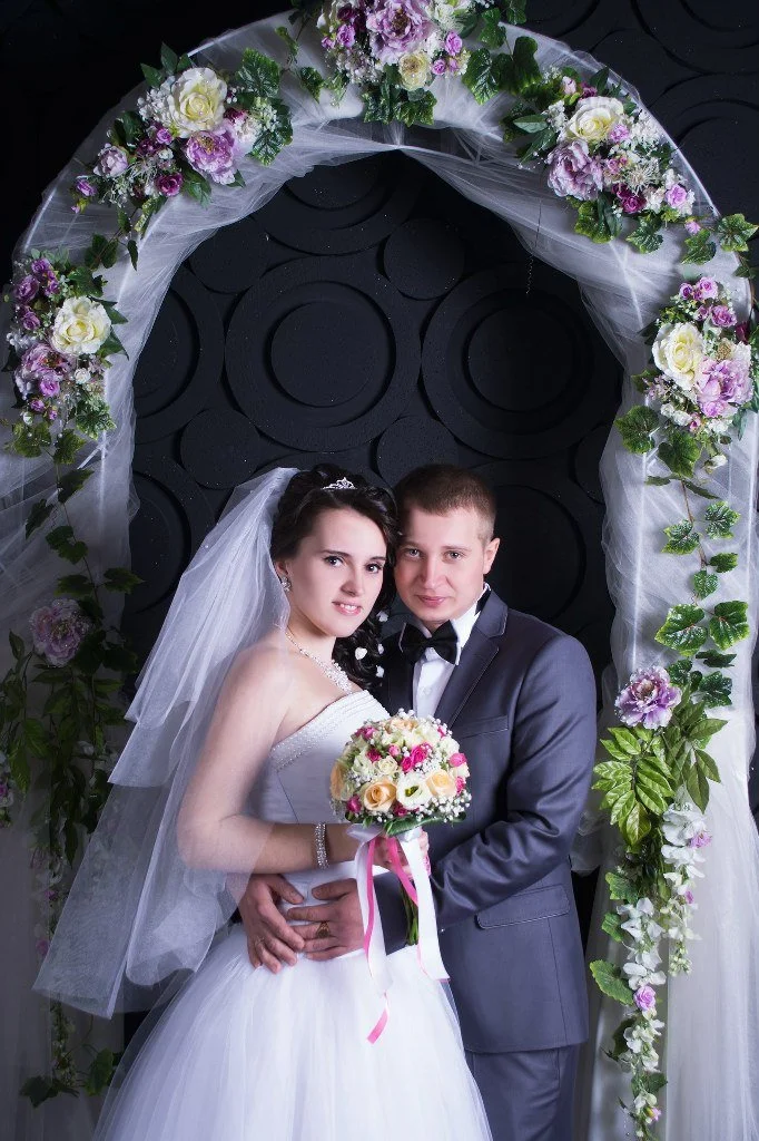 A bride and groom standing under a floral wedding arch, with the bride holding a bouquet of flowers.