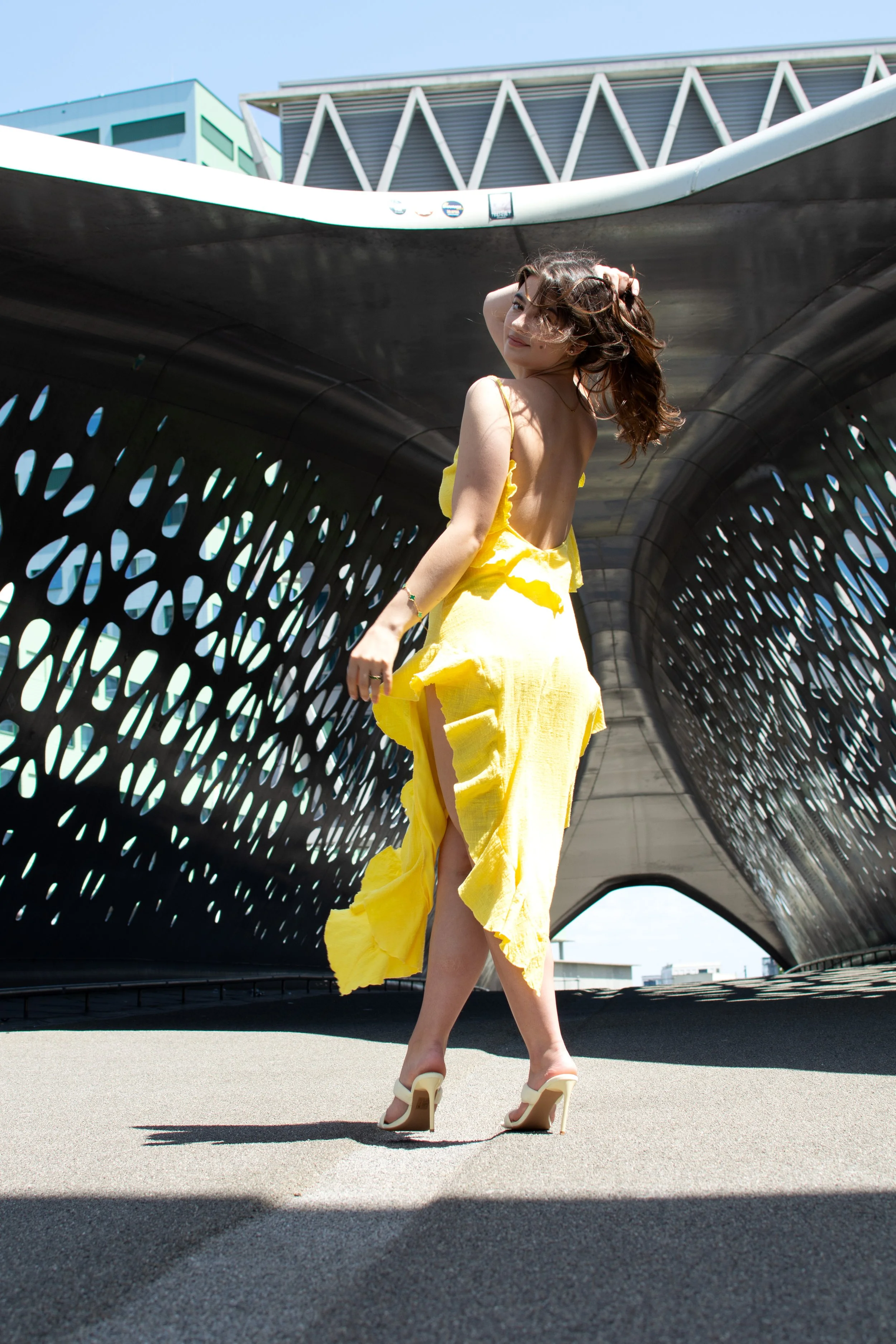 A woman in a yellow dress and high heels standing in front of a modern architectural structure on a sunny day.