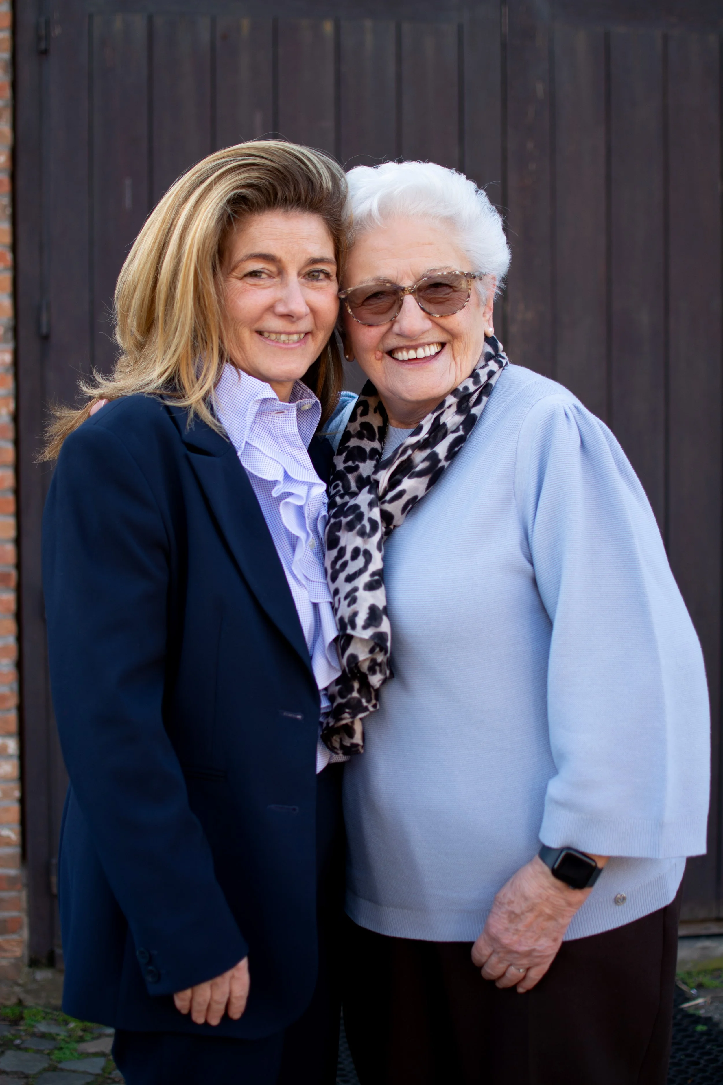 Two women standing outdoors, embracing and smiling at the camera, with a wooden door and brick wall in the background.