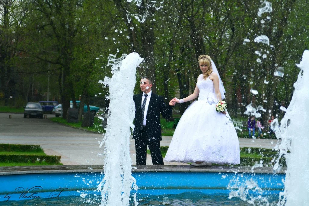 A bride and groom holding hands by a fountain in a park, with water splashing around them.