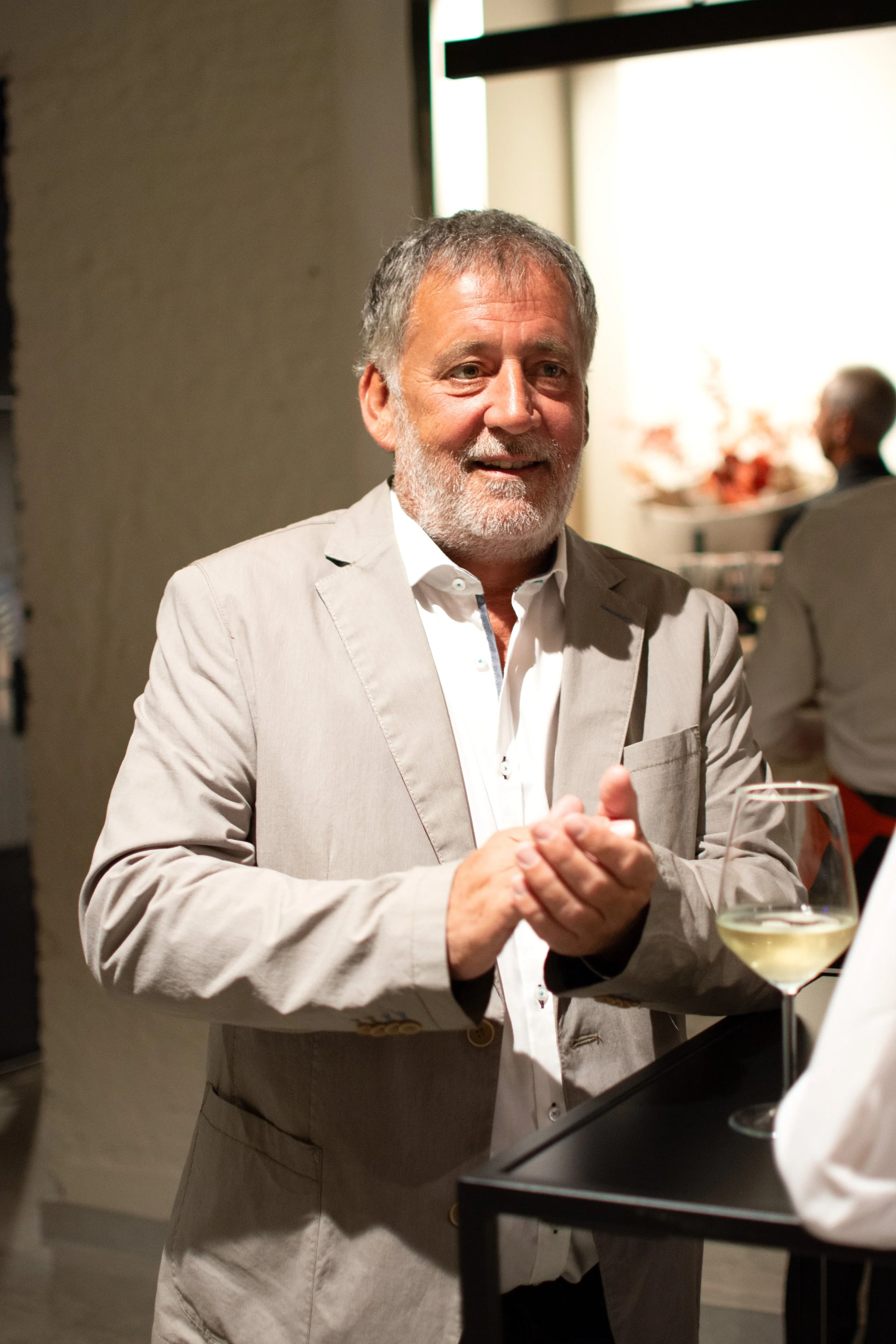 A smiling older man with gray hair and beard, wearing a light gray suit and white shirt, standing at a bar or counter with a glass of white wine in front of him, in a well-lit indoor setting.