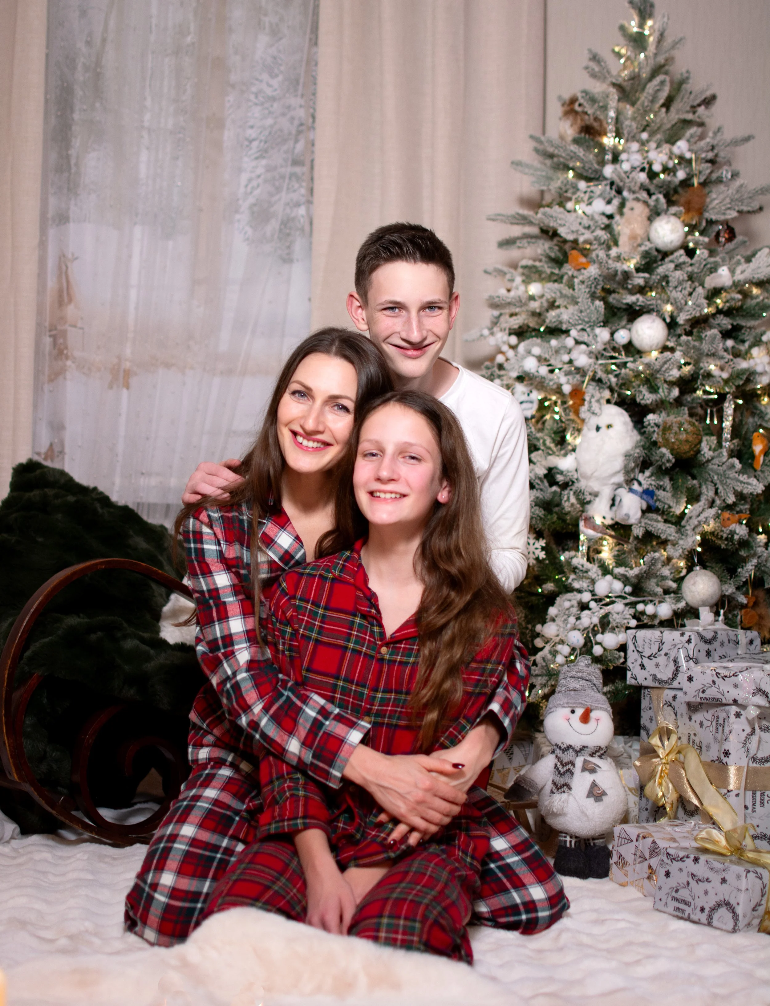 A woman, a teenage boy, and a young girl posing together in front of a decorated Christmas tree with wrapped gifts and a snowman plush toy in the background.