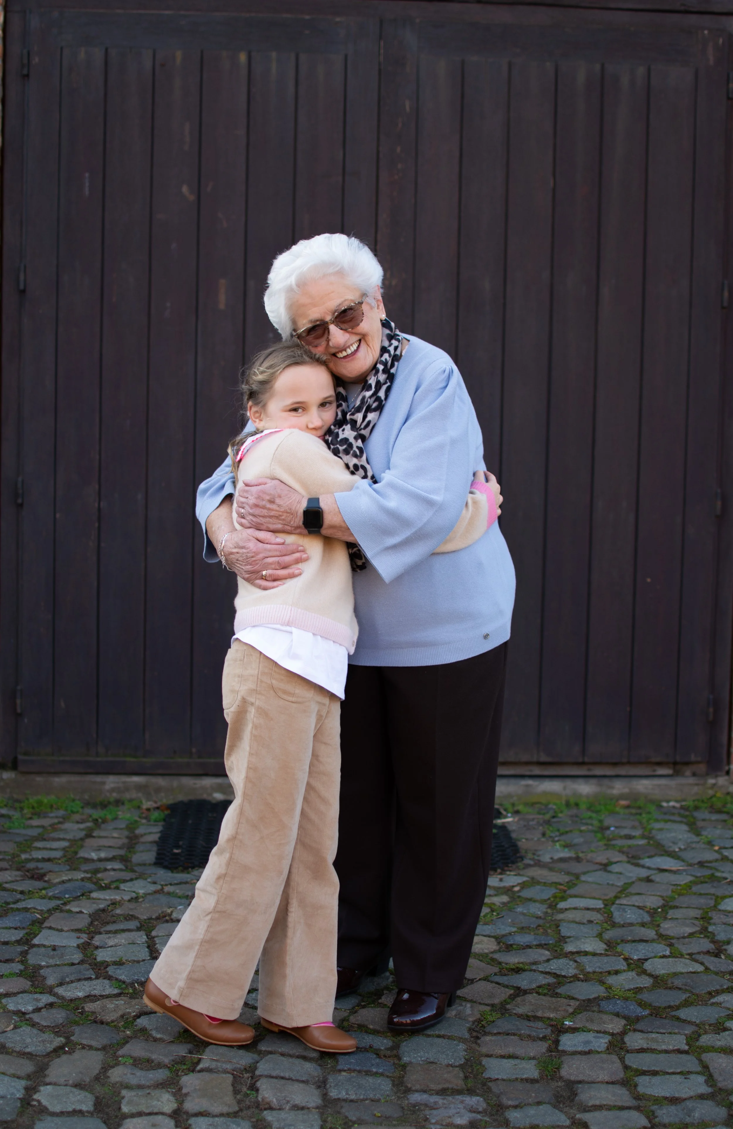 An elderly woman with white hair and glasses hugging a young girl outside, standing on a cobblestone area in front of a dark wooden fence, both smiling.