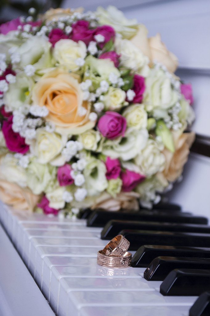 Wedding rings placed on a piano keyboard with a bouquet of white, pink, and cream roses in the background.
