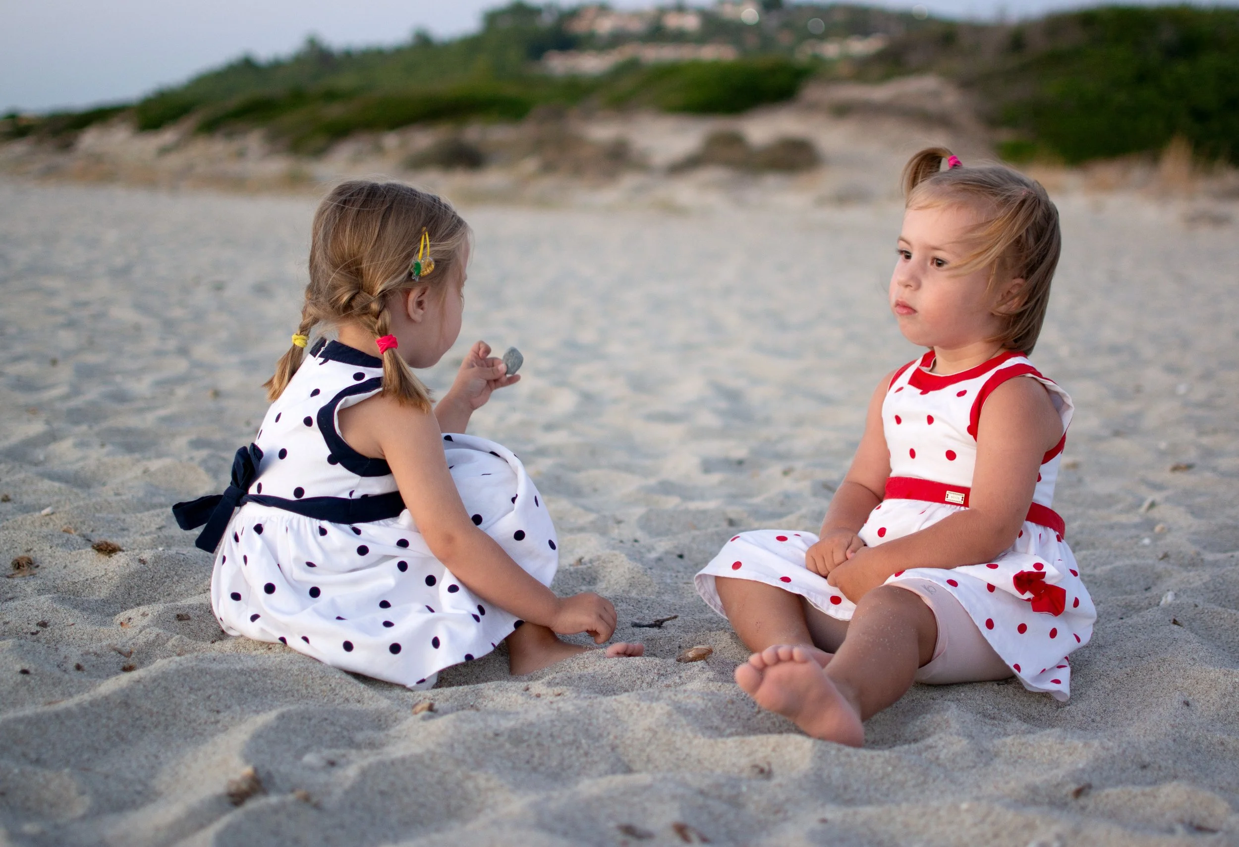 Two young girls sitting on the sandy beach, one holding a small object, with a hilly landscape and vegetation in the background, dressed in white dresses with polka dots.