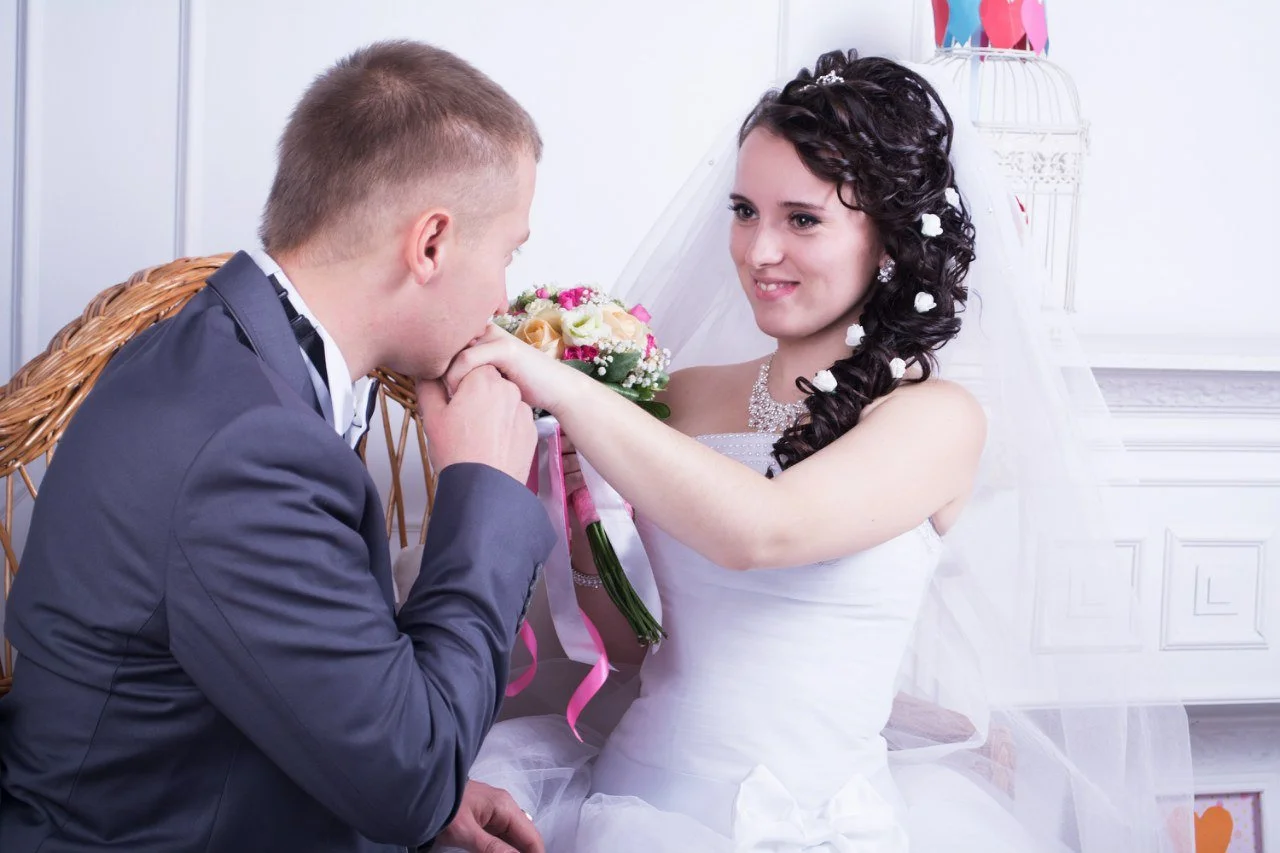 A bride holding a bouquet of flowers smiles as a groom, kneeling, kisses her hand during a wedding ceremony.