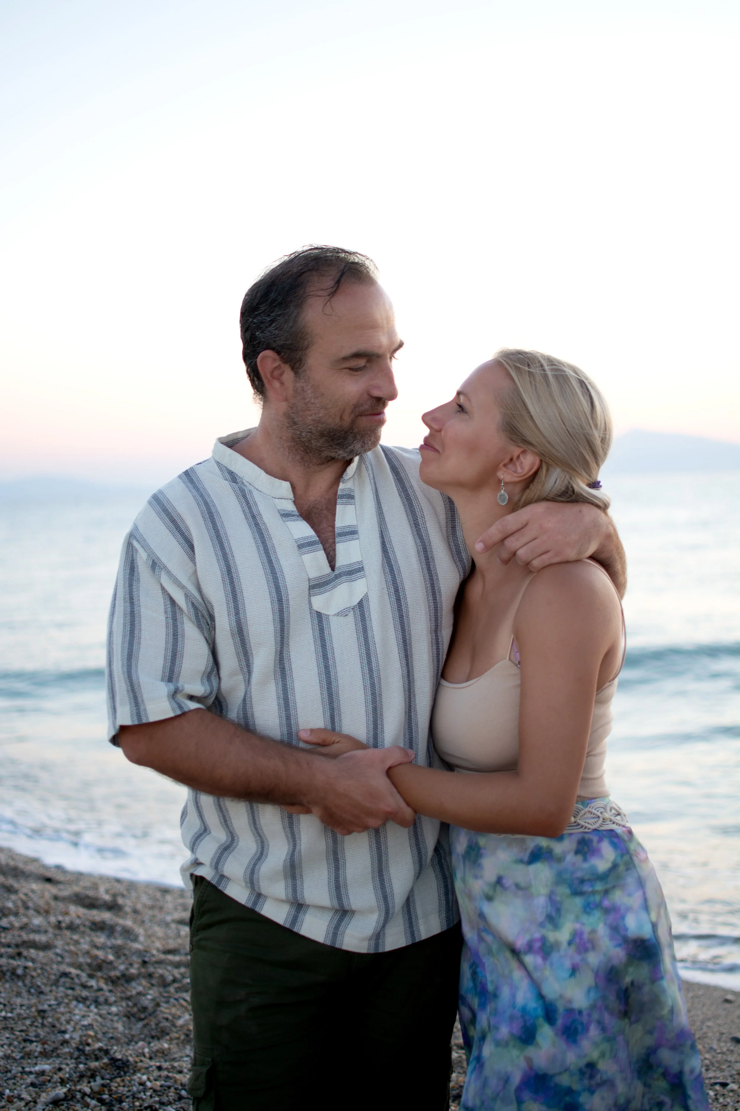 A romantic couple stands on a beach at sunset, embracing each other and looking into each other's eyes.