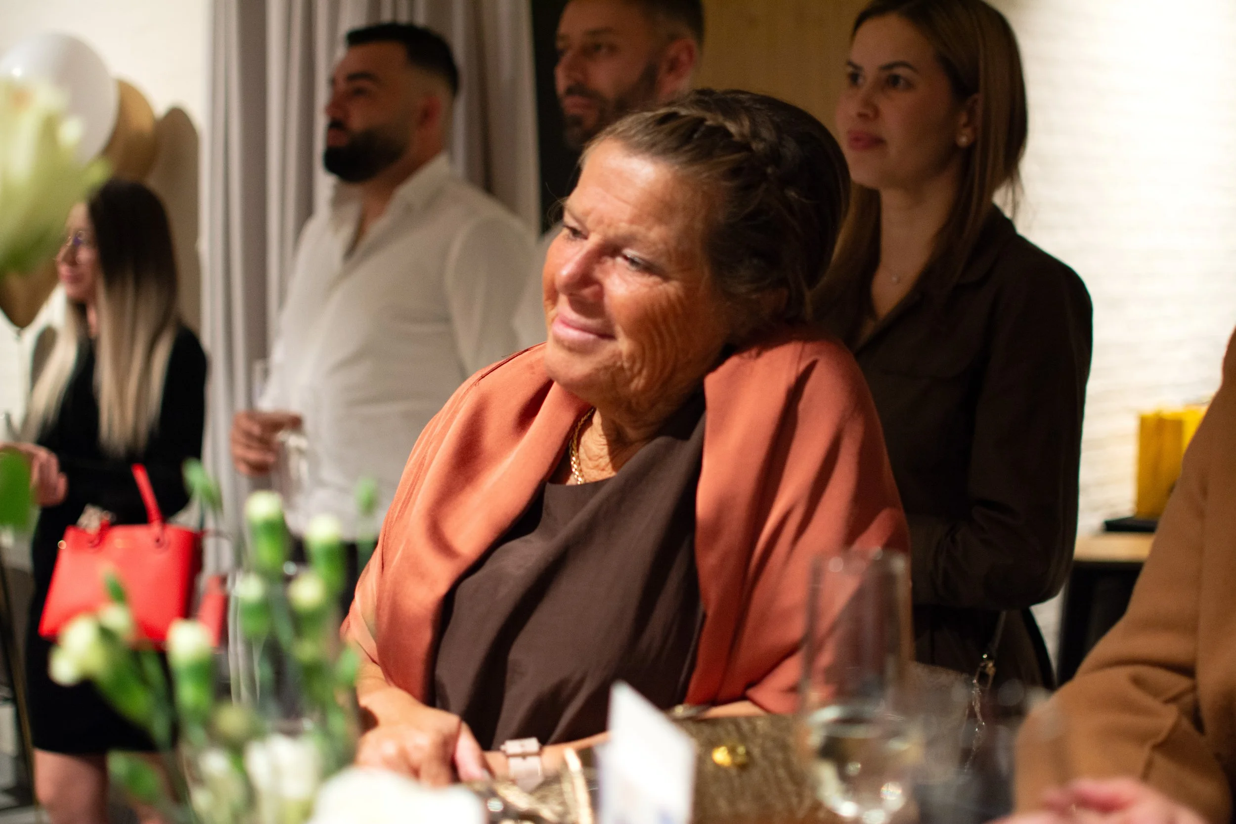 An elderly woman with short, dark hair styled back, wearing a brown and peach shawl, smiling and leaning her head on a woman's shoulder in an indoor setting with other people in the background.