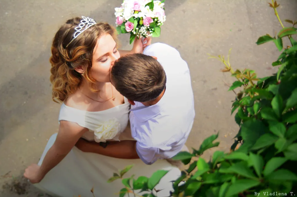 Overhead view of a couple kissing, the woman wearing a white dress and a tiara, holding a bouquet of pink and white flowers, with green foliage in the foreground.