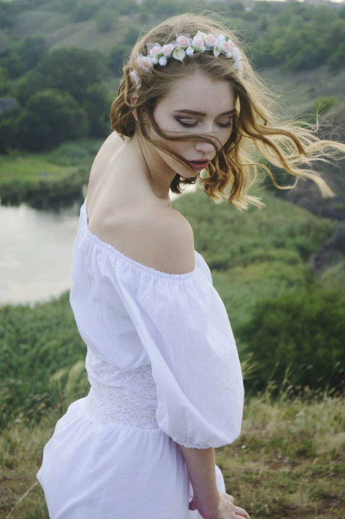 A young woman with wavy, shoulder-length red hair, wearing a white off-the-shoulder dress and a floral headband, standing outdoors near a body of water with green hills in the background.