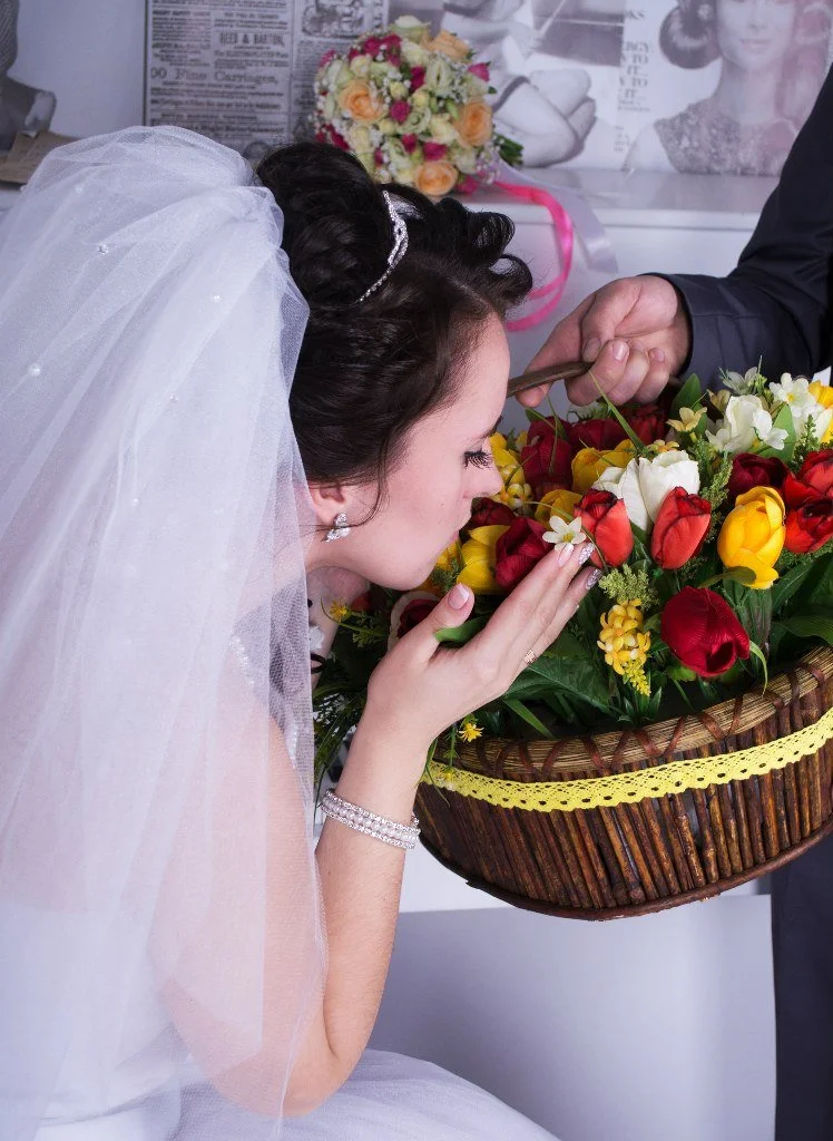 Bride in wedding dress and veil closes her eyes and gently kisses a bouquet of colorful flowers, as an arm extends a hand holding a knife for a flower-gifting ceremony.