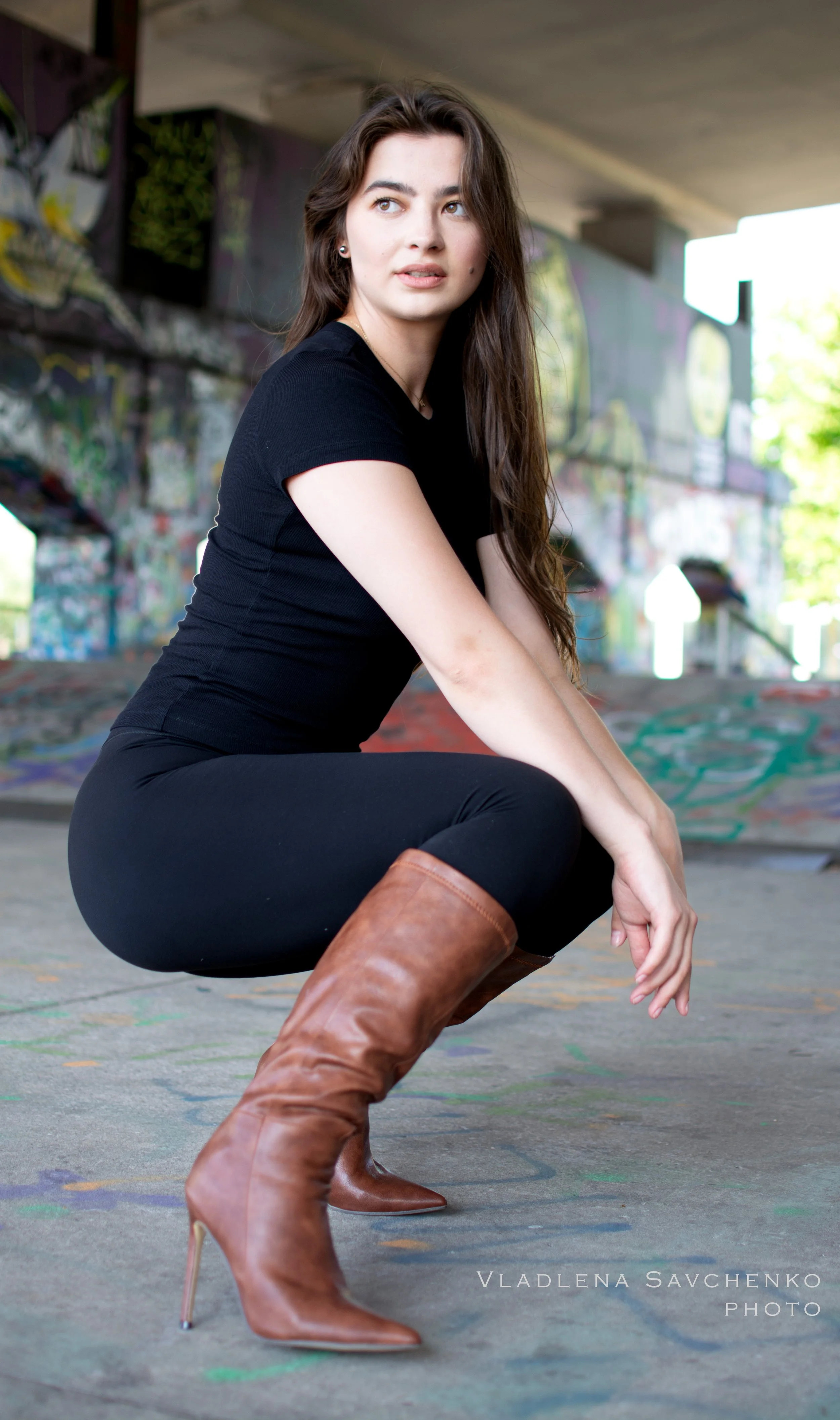 A woman with long brown hair, wearing a black shirt, black pants, and brown high-heeled boots, squatting under a graffiti-covered bridge.