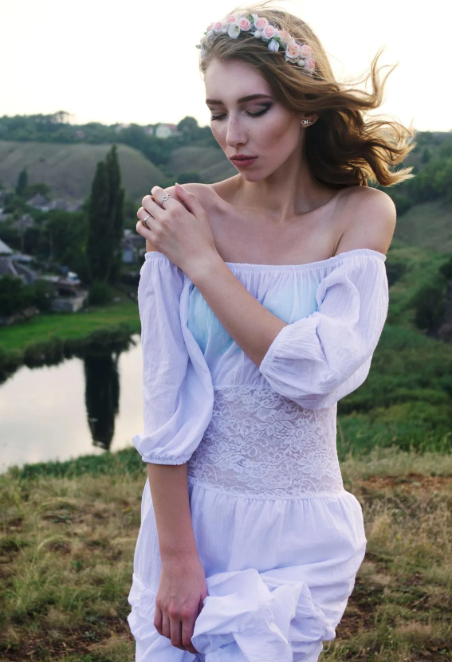 A young woman in a white off-the-shoulder dress with lace details, wearing a flower crown, standing outdoors near a river with hills and trees in the background.