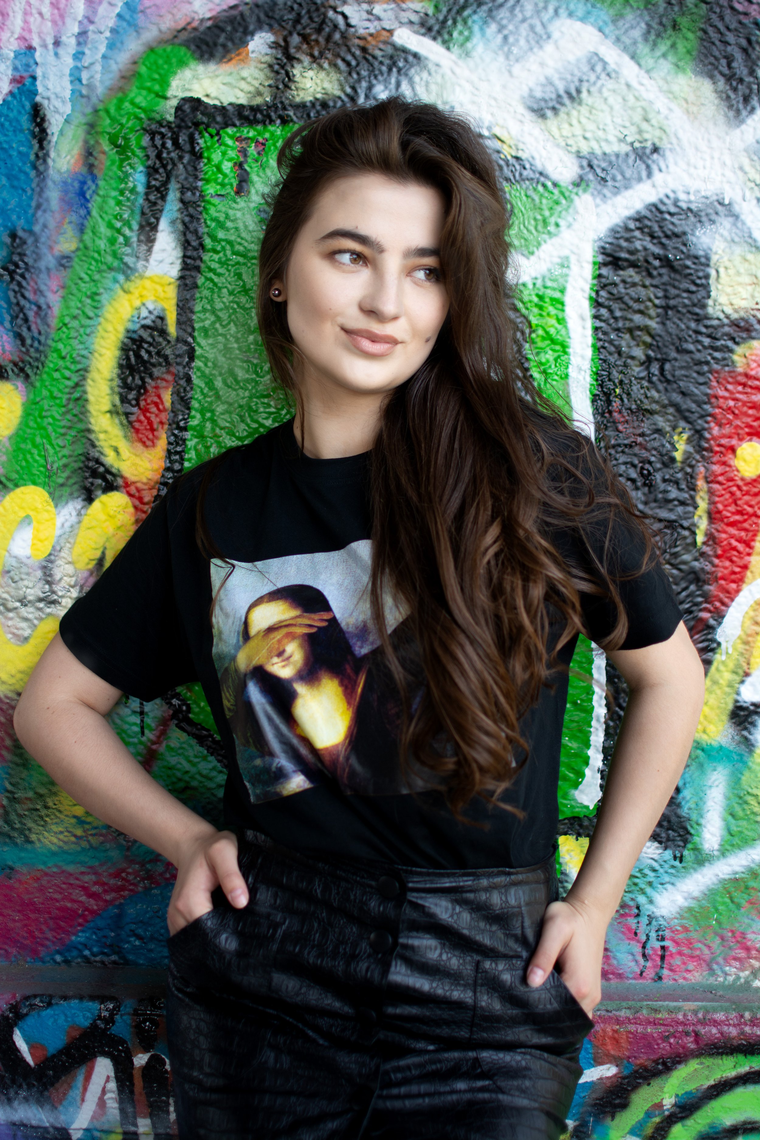 A young woman with long wavy brown hair wearing a black t-shirt with an image of the Mona Lisa and black textured pants, standing in front of a colorful graffiti wall.