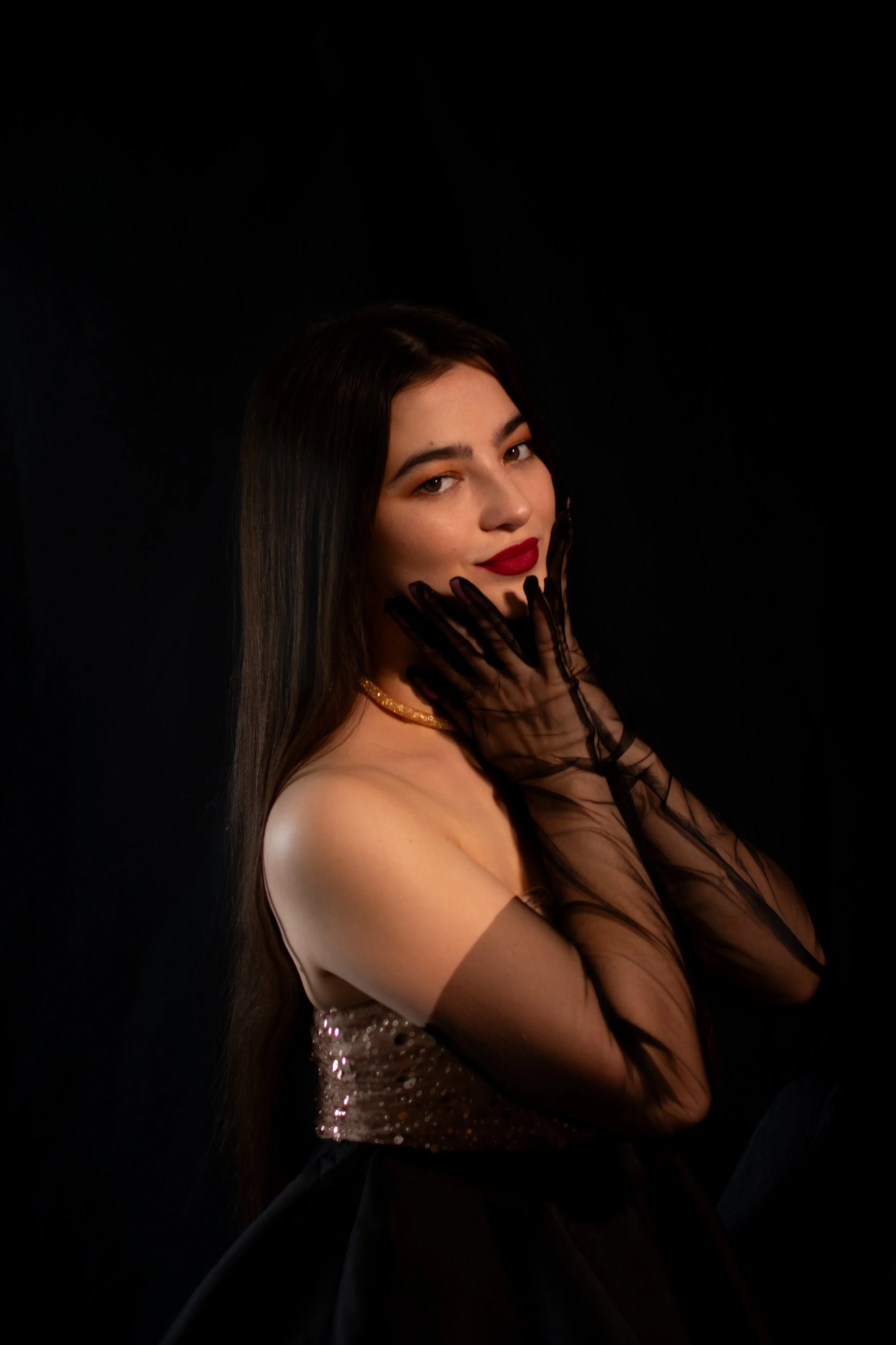 A young woman with long dark hair, red lipstick, and gold jewelry poses against a black background. She wears sheer black gloves and a sequined strapless dress, with her hand gently touching her face.