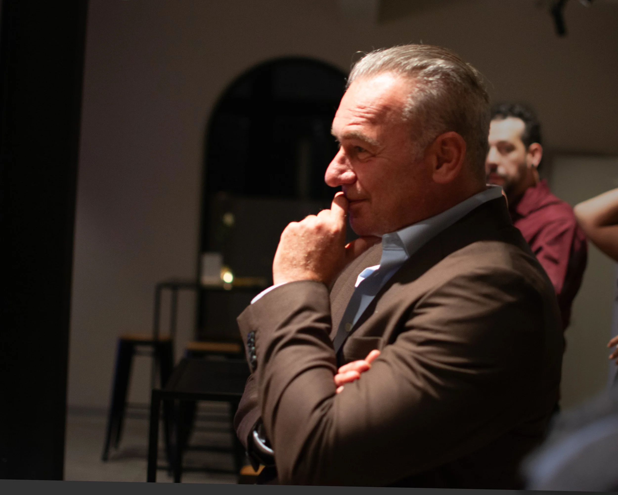 A man in a brown jacket with a blue shirt, sitting with his hand on his chin, appearing thoughtful, with another man in a red shirt blurred in the background.