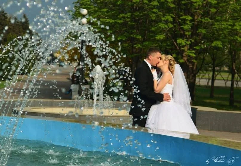 A bride and groom sharing a kiss by a fountain with water splashing in the foreground, outdoors with green trees in the background.