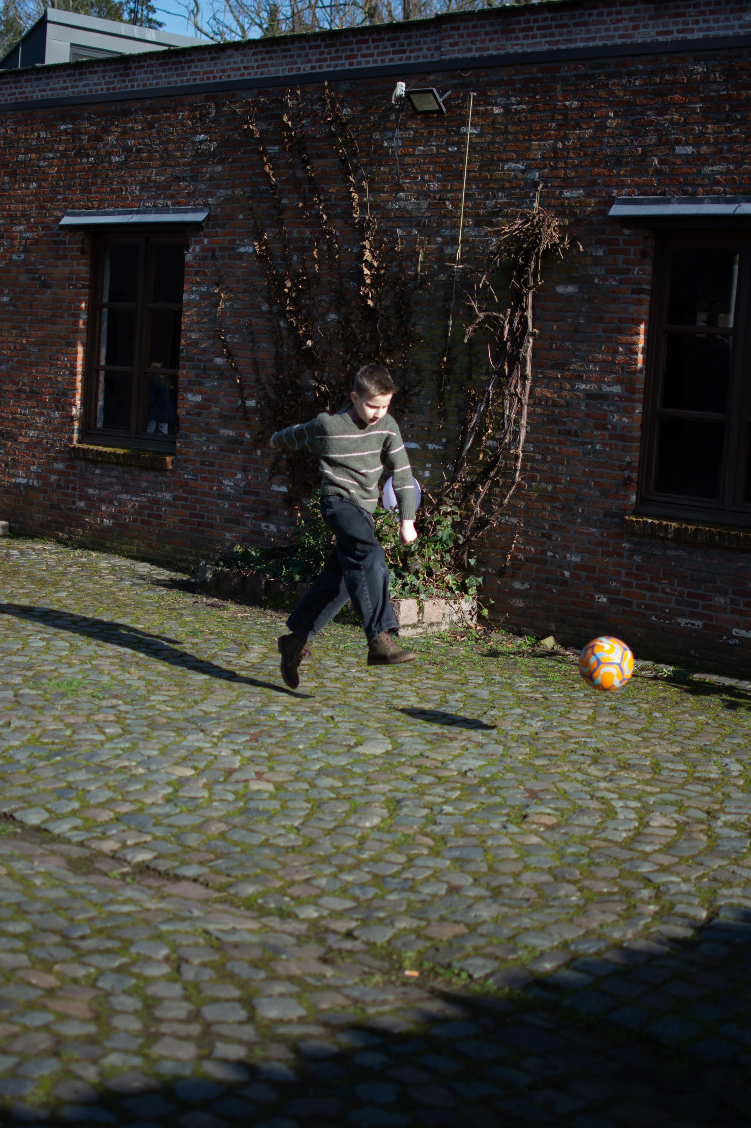 A boy playing soccer on a cobblestone street, kicking an orange and white ball in front of a brick building with two black-framed windows.