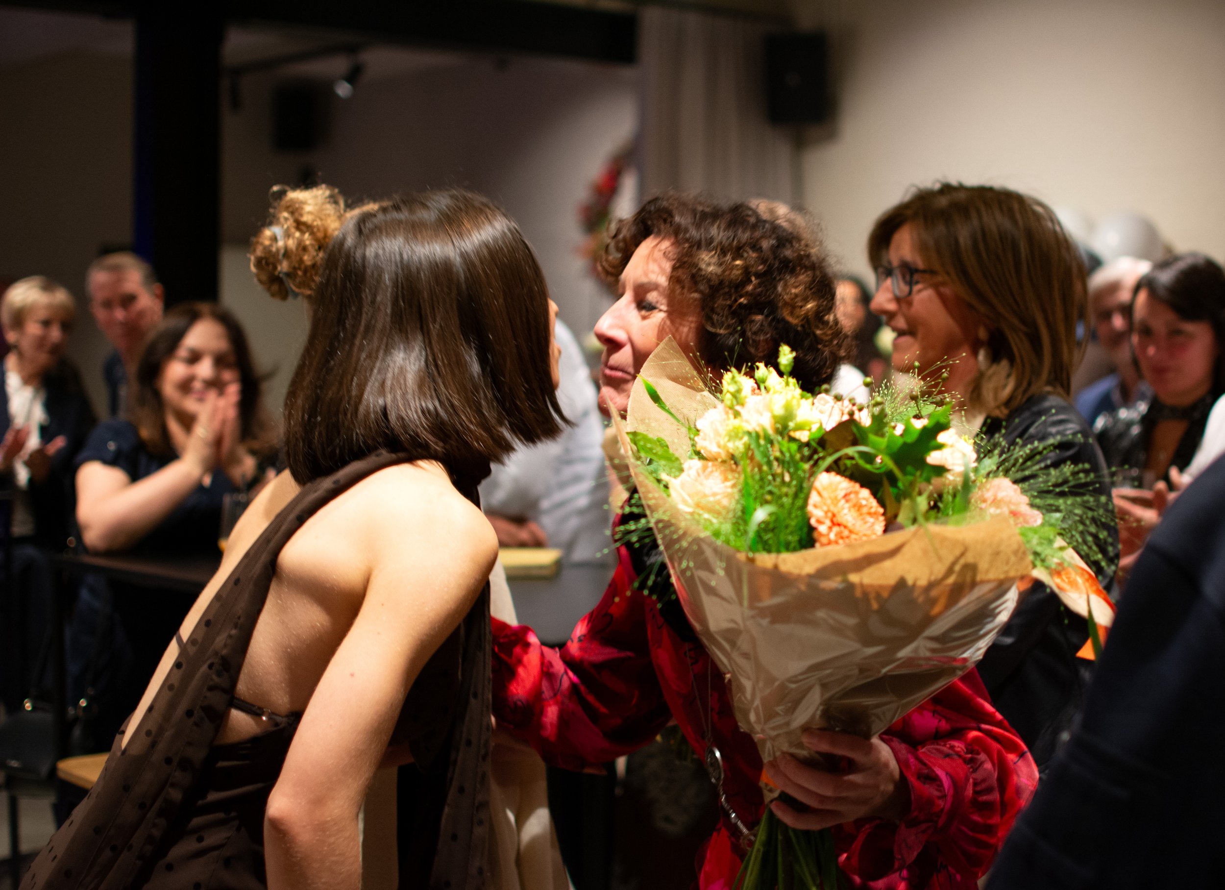 A woman in a red dress is holding a bouquet of flowers and hugging a young girl with shoulder-length brown hair, while others smile and clap in the background at an indoor event.