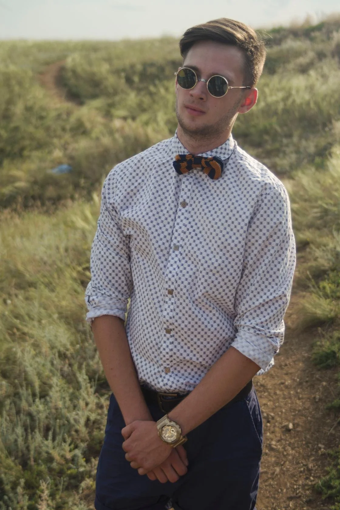 A young man with dark hair, wearing round sunglasses, a patterned white button-up shirt with rolled-up sleeves, a leopard-print bow tie, and a gold watch, standing outdoors in a grassy field.