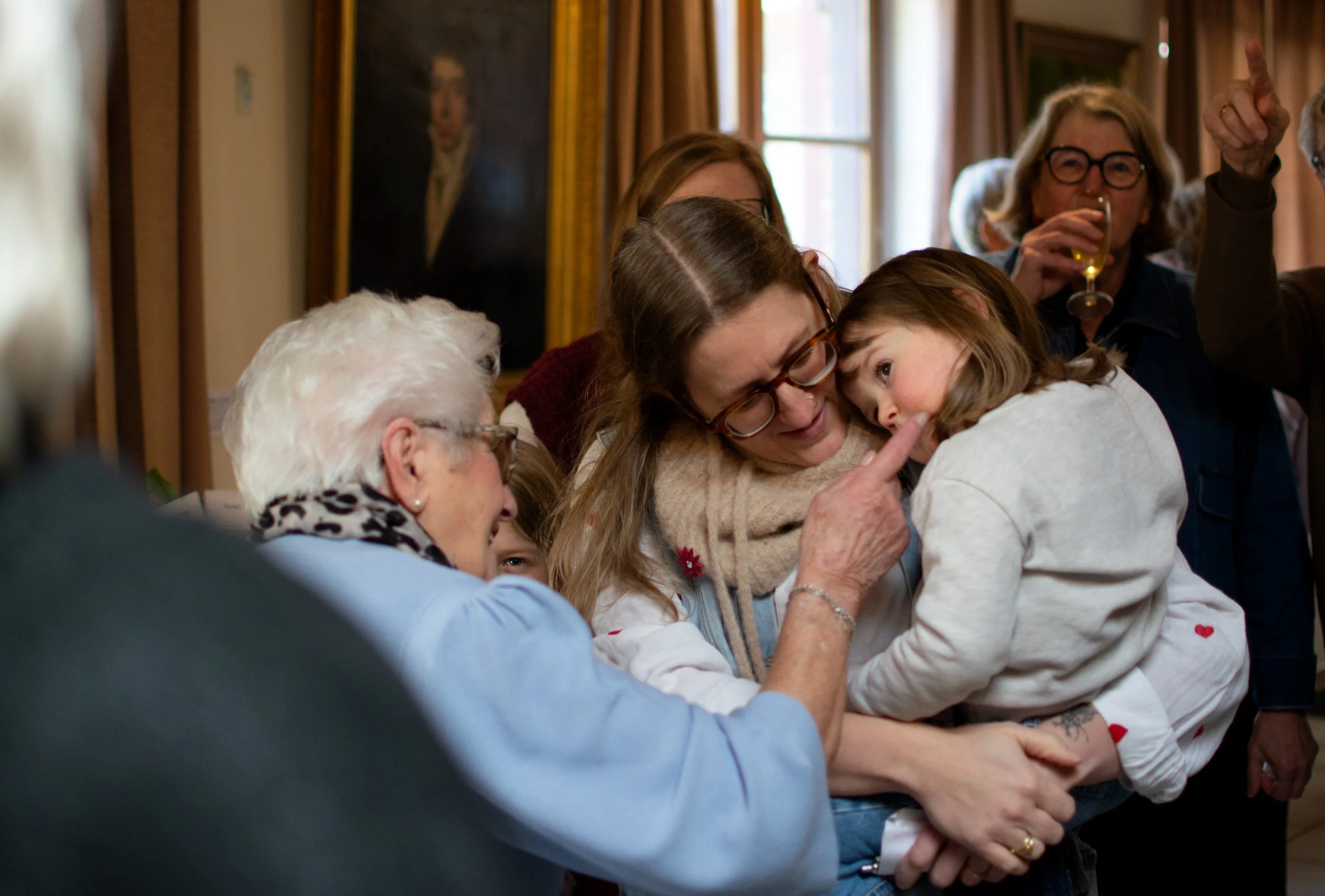 Multiple people, including children and elderly, sharing a warm moment together indoors, with some smiling and hugging.
