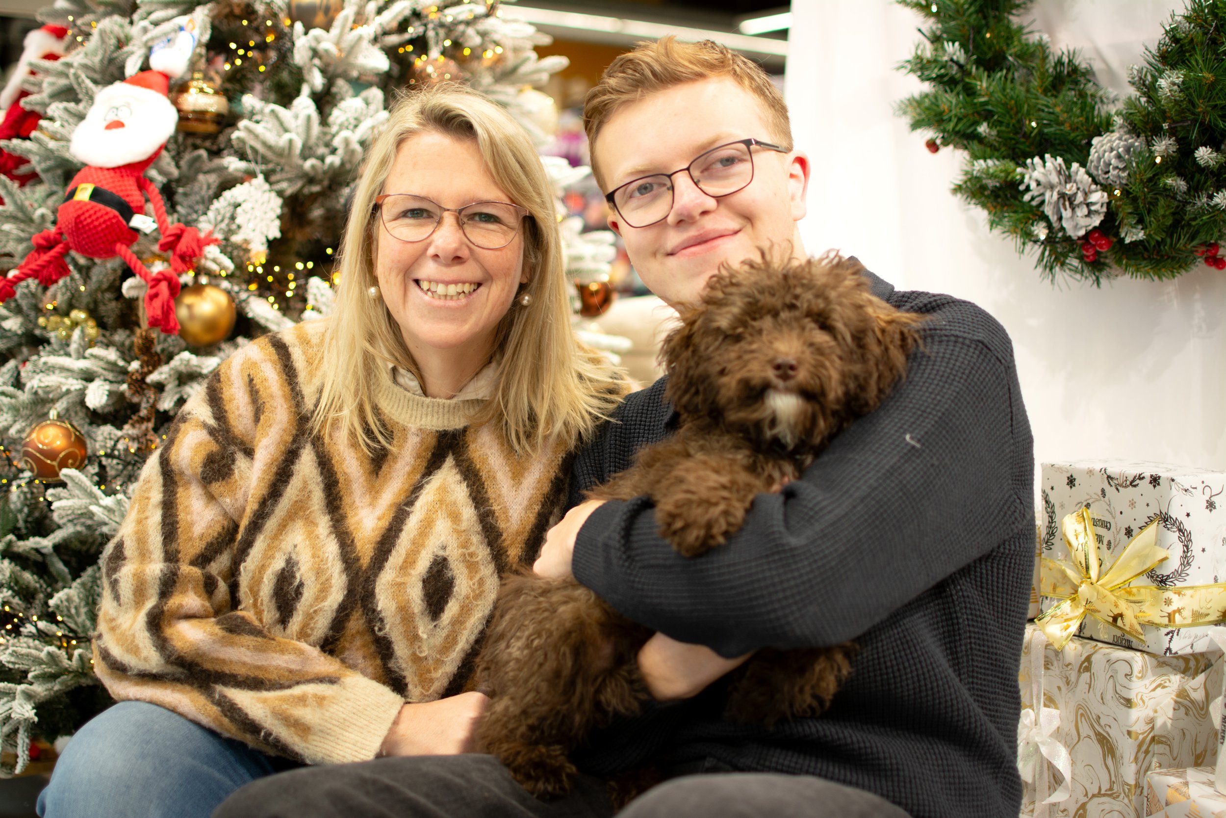 A smiling woman with blonde hair and glasses, a young man with glasses, and a brown fluffy dog in front of Christmas trees with ornaments and gifts.