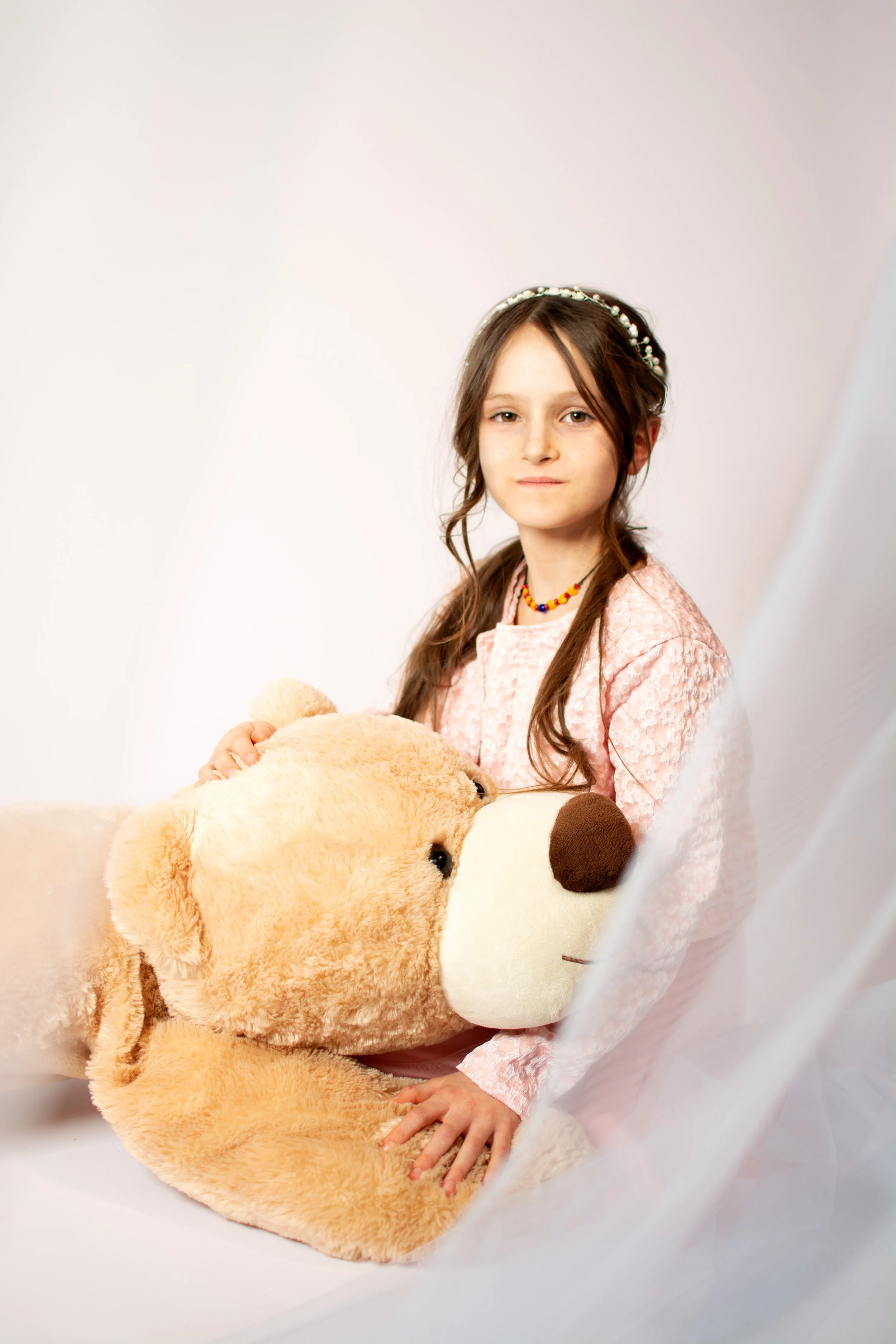 A young girl with long brown hair, wearing a pink dress and a beaded necklace, sits with a large stuffed teddy bear against a light background.