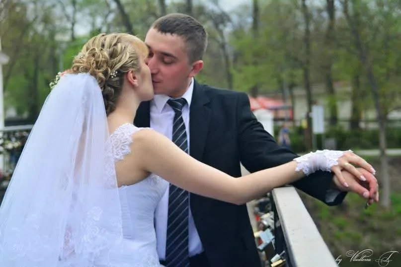 A bride and groom sharing a kiss outdoors, with the bride wearing a white wedding dress and veil, and the groom in a black suit and tie, standing near a railing.