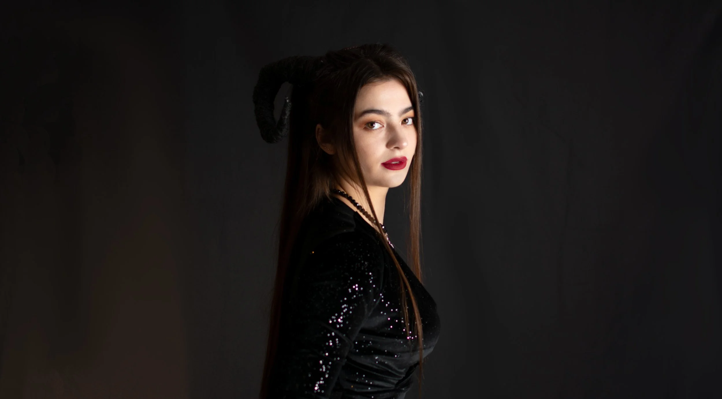 A woman with long brown hair, red lipstick, and dark eye makeup wearing a black, sequined long-sleeve top and necklace, posing against a black background with horns on her head.
