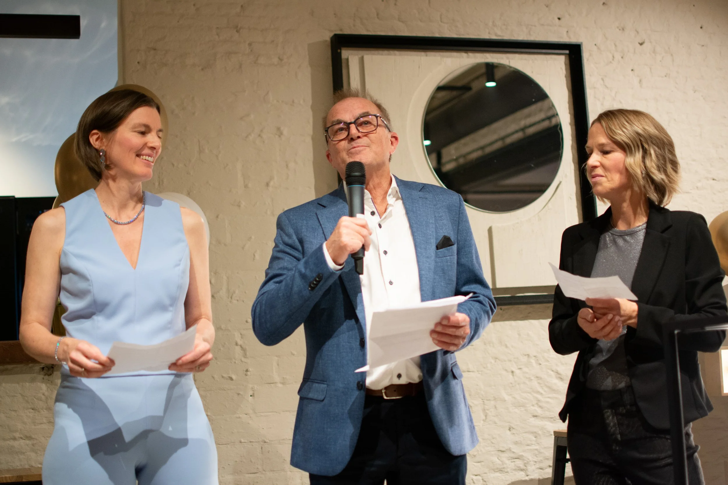 Three people standing together at a formal event, with a man in the middle holding a microphone, wearing a blue blazer, and the two women on either side holding papers. They are in front of a white brick wall and a circular mirror.