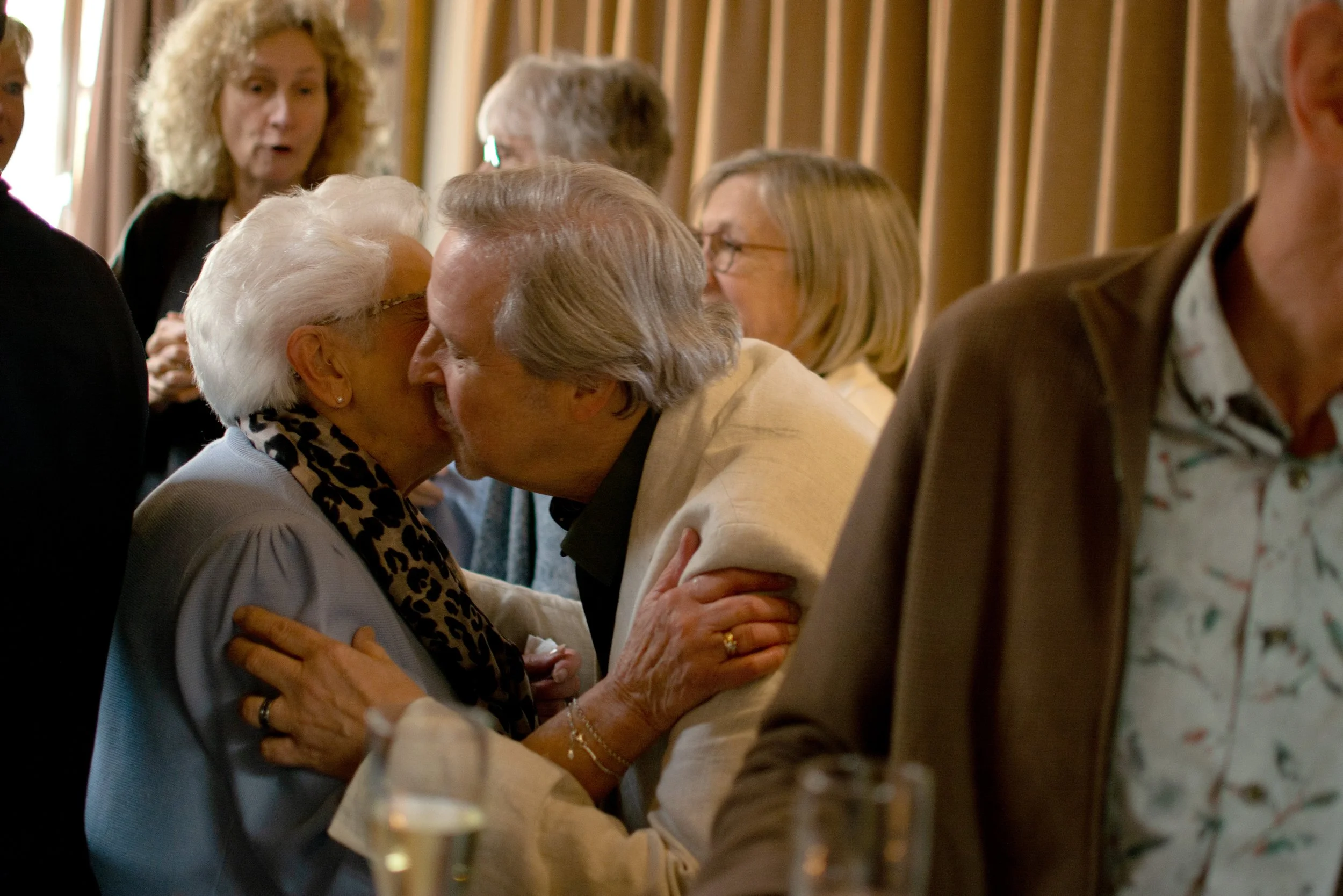 Two elderly women are greeting each other with a kiss on the cheek at a social gathering, surrounded by other people in a warmly lit room.