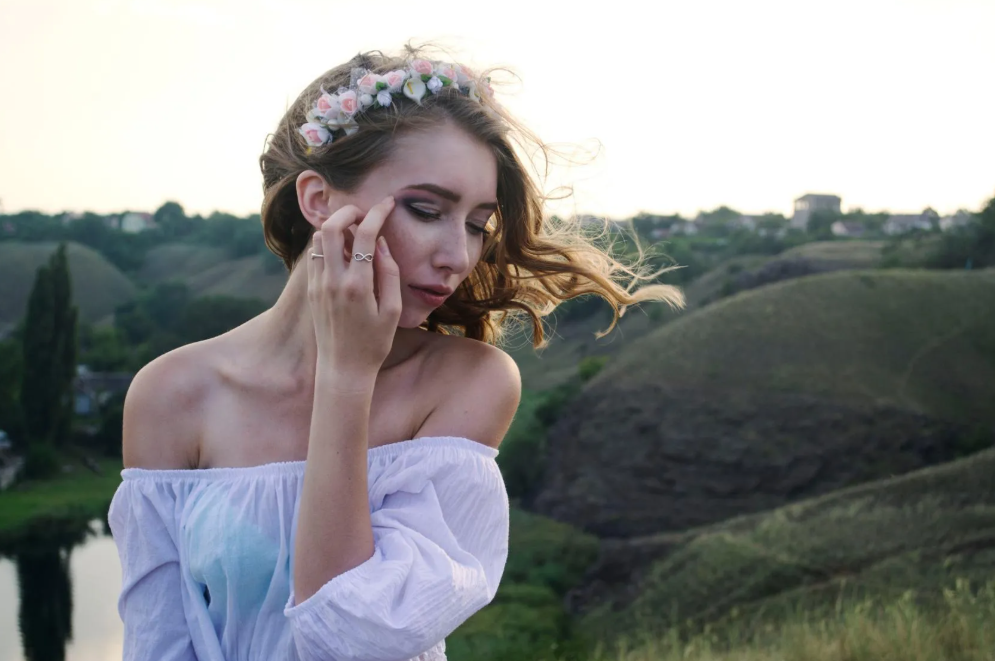 Young woman with long flowing hair wearing a flower crown and off-the-shoulder white dress, standing outdoors near a river with green rolling hills in the background.
