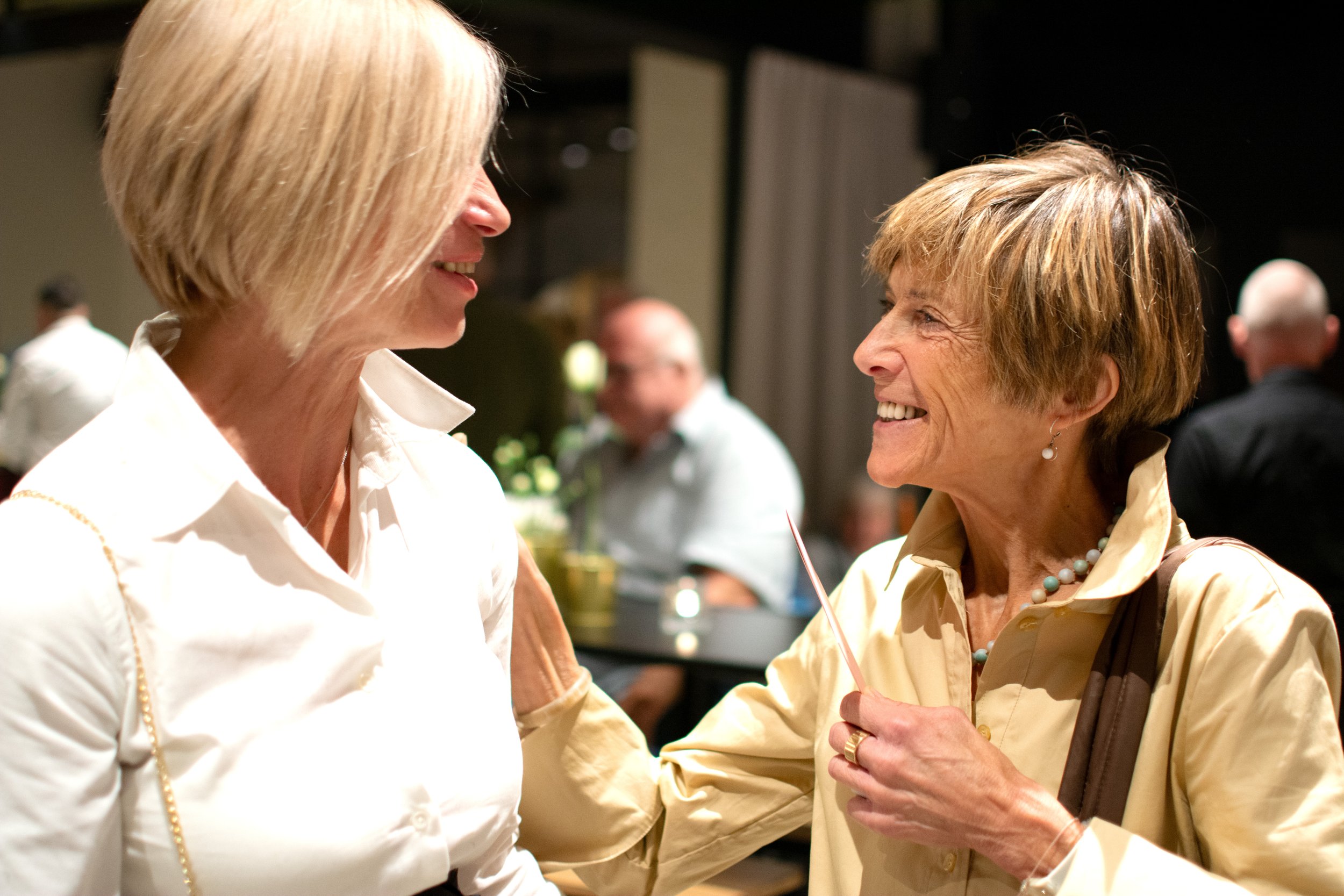 Two women smiling and talking at a social gathering, with other people in the background.