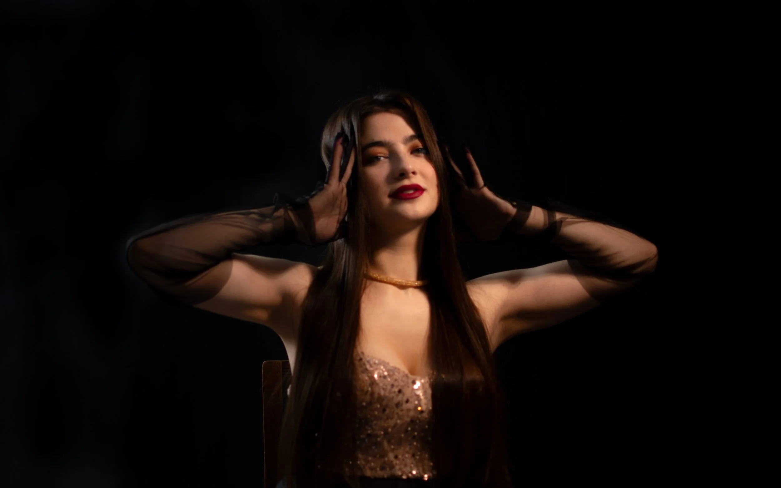 A woman with long dark hair and red lipstick posing against a black background, wearing sheer black gloves and a shiny, sequined, bronze-colored top.