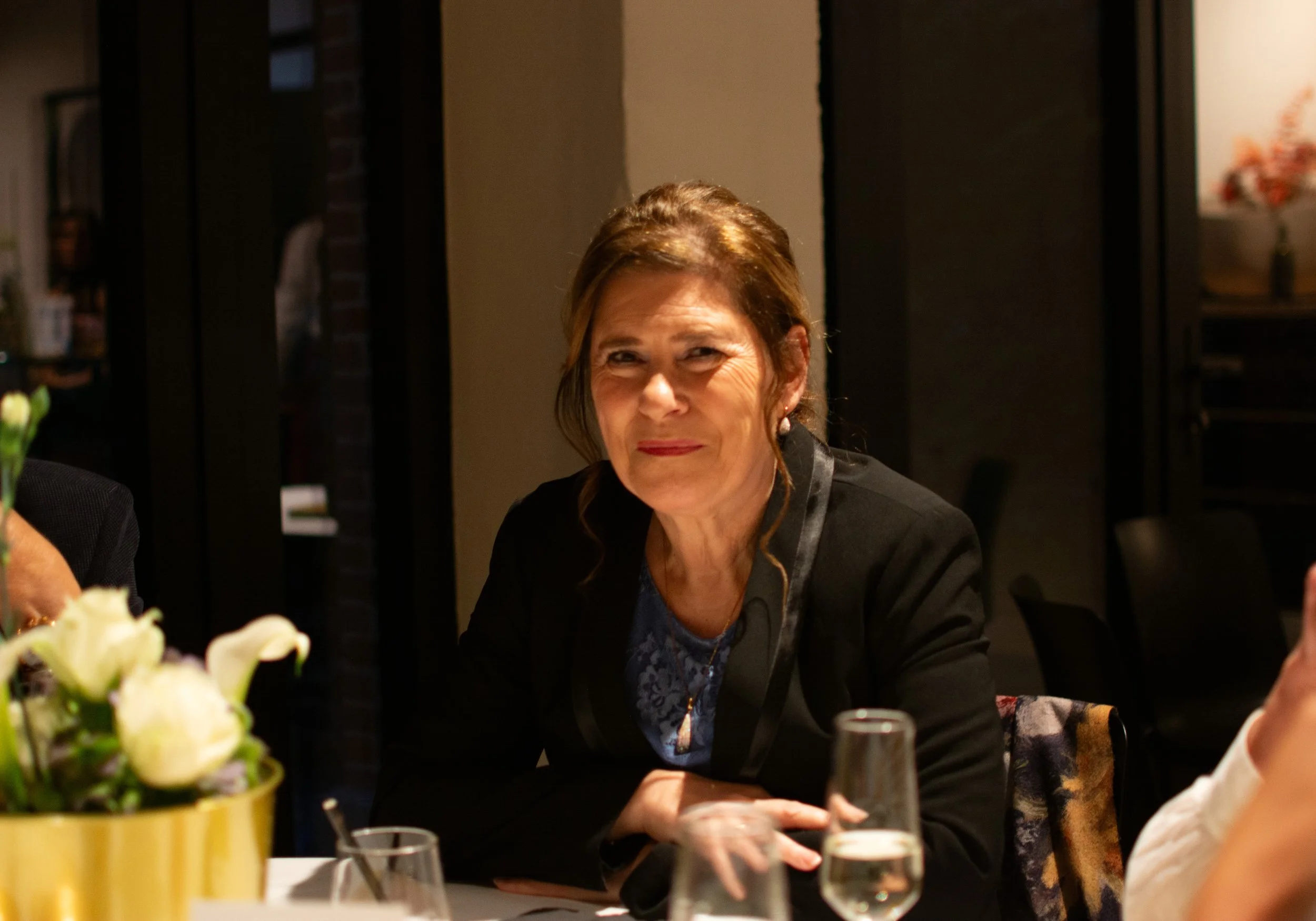 A middle-aged woman with light skin and brown hair, smiling, sitting at a dinner table during an indoor event with wine glasses and flowers.