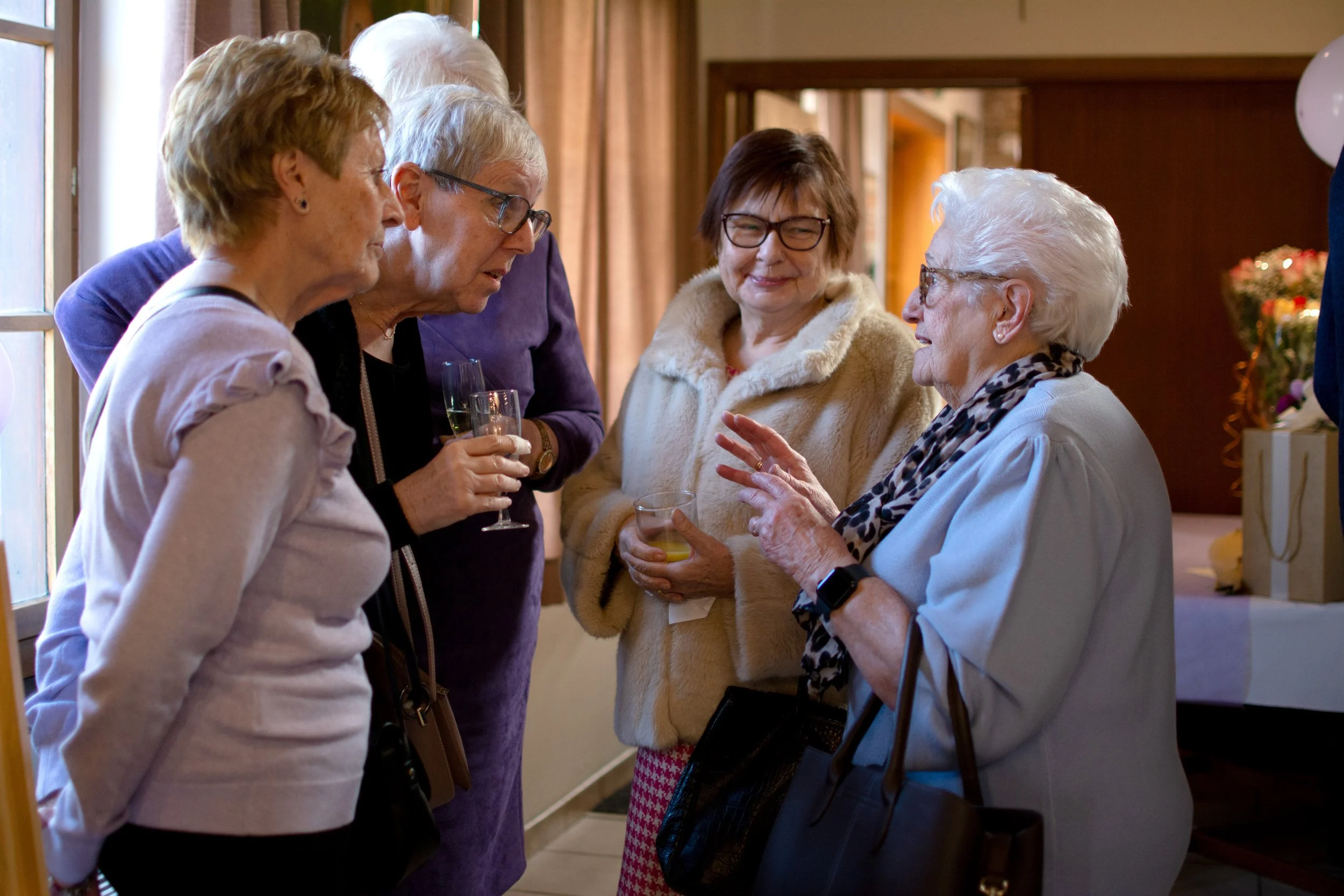 Group of elderly women engaged in conversation at a social gathering, holding drinks, indoors by a window.