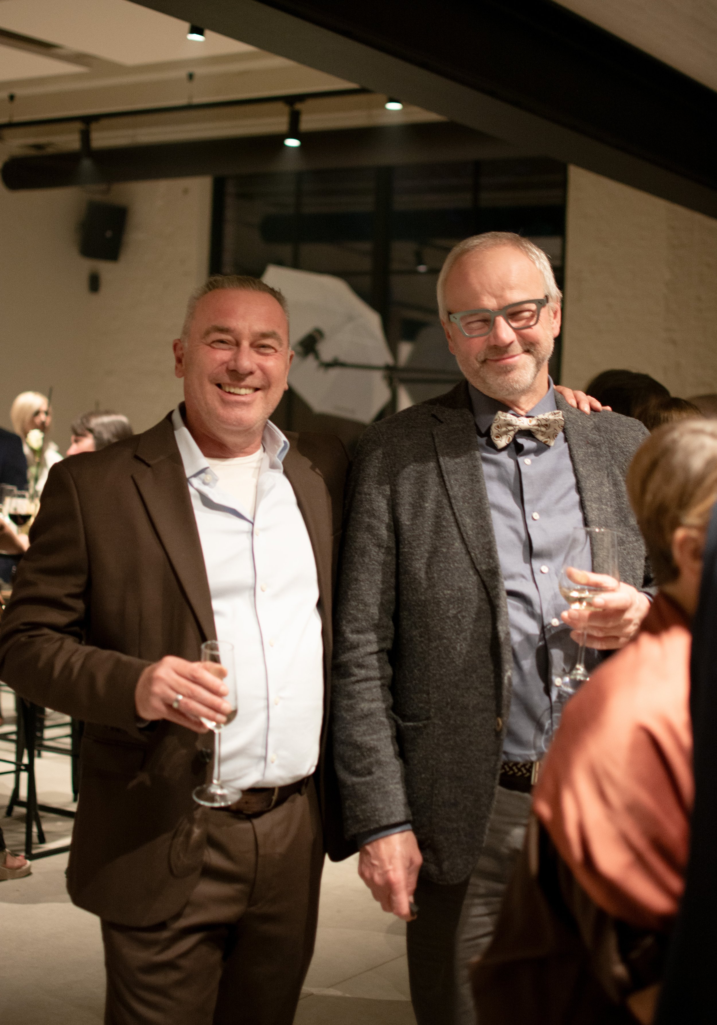 Two men smiling and holding glasses of wine at an indoor event or party.