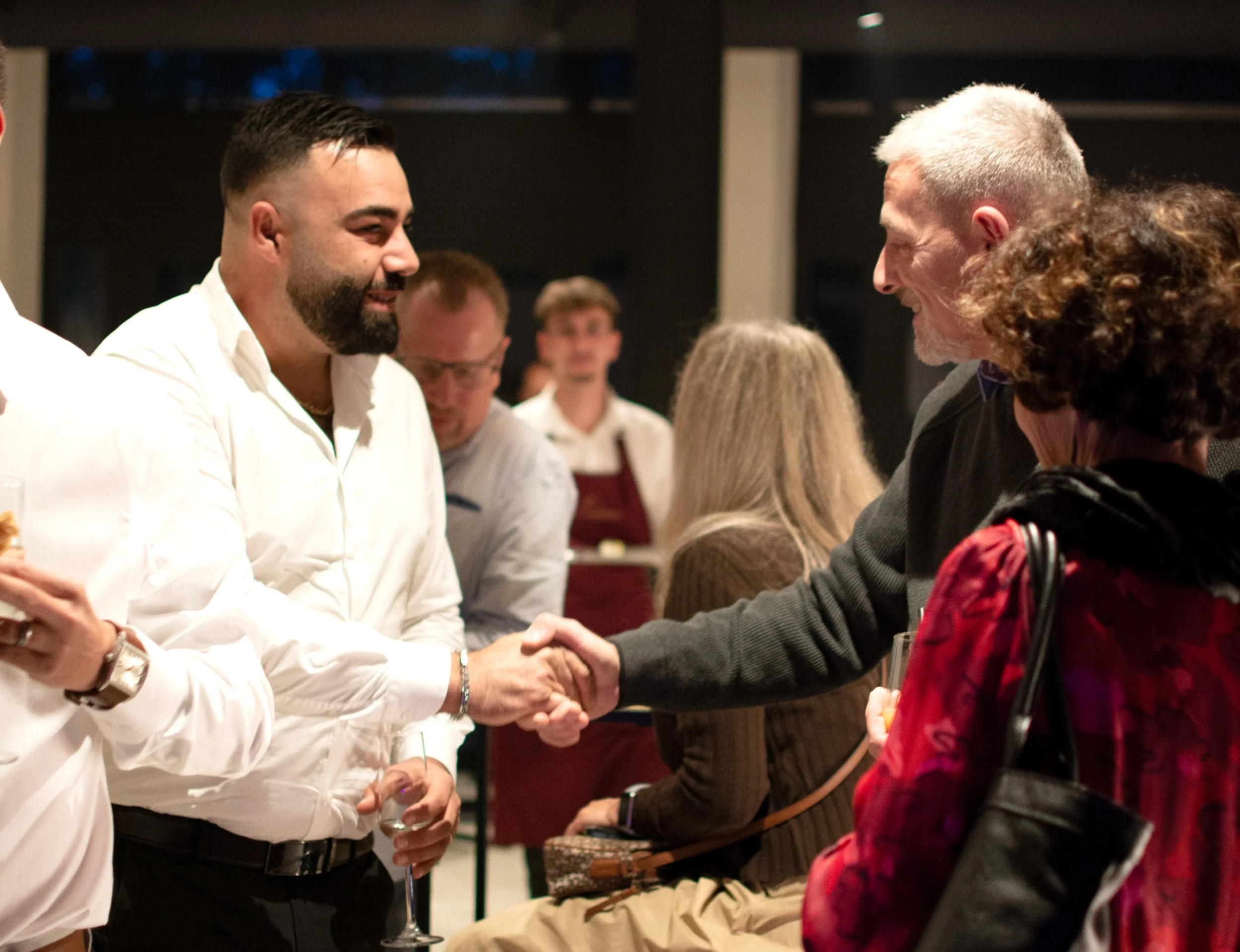 A man with a beard and white shirt shaking hands with an older man with white hair and beard, at a social gathering or reception.