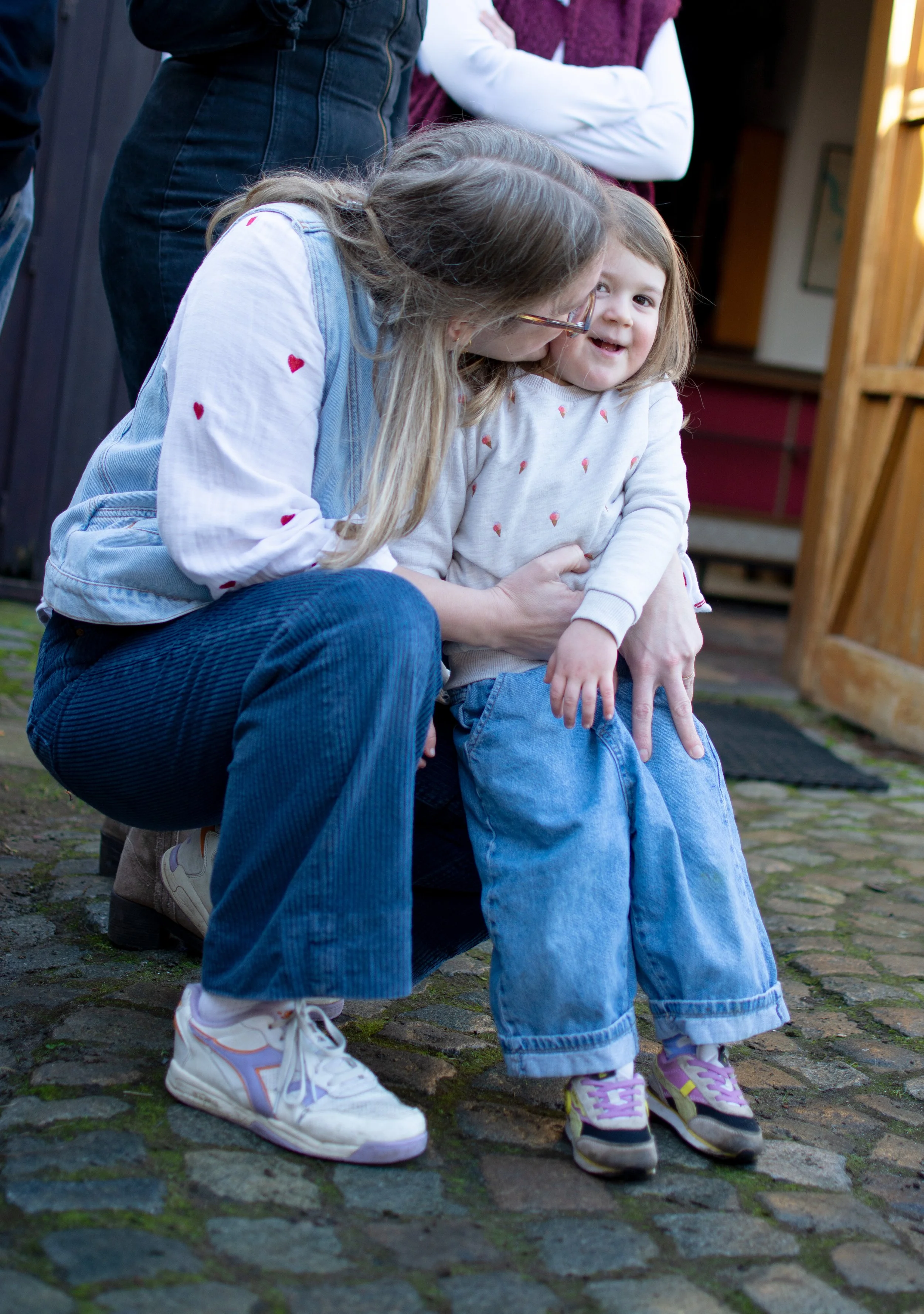A woman crouching down and smiling at a young girl, possibly her daughter, who is also smiling and looking happy. The woman is wearing glasses, a light blue vest, and dark blue pants. The girl is wearing a white sweatshirt with small red hearts, jean