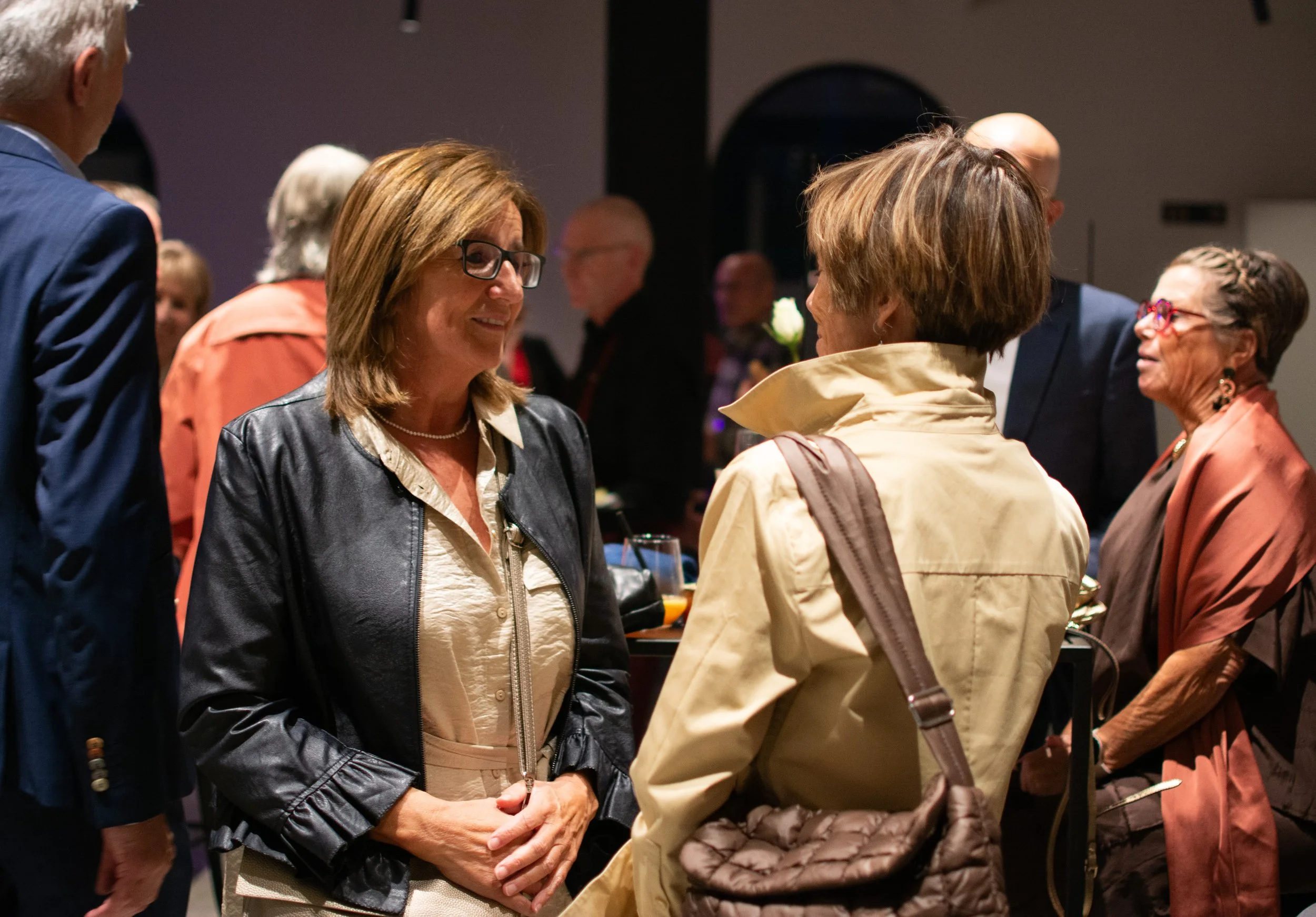 Group of people socializing, two women in the foreground engaged in conversation at an indoor social event.