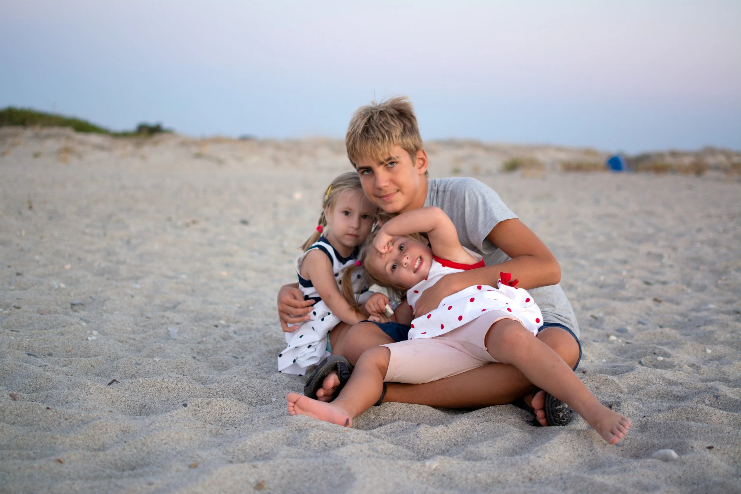 A teenage boy with two young girls sitting on the sandy beach with a distant horizon and sky in the background.