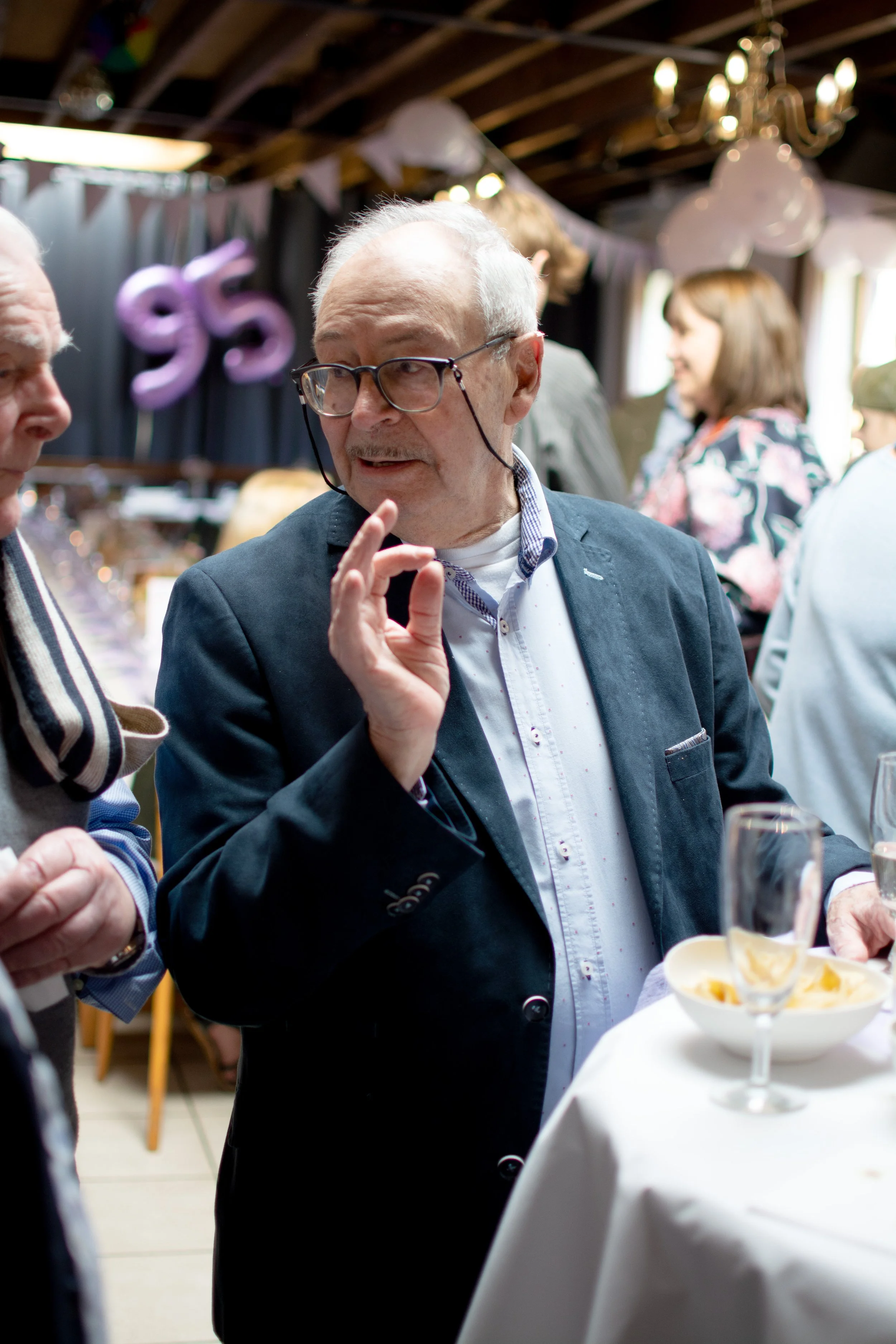 An elderly man in glasses and a dark blazer gestures while talking to another person at an indoor celebration, with balloons and a chandelier in the background.